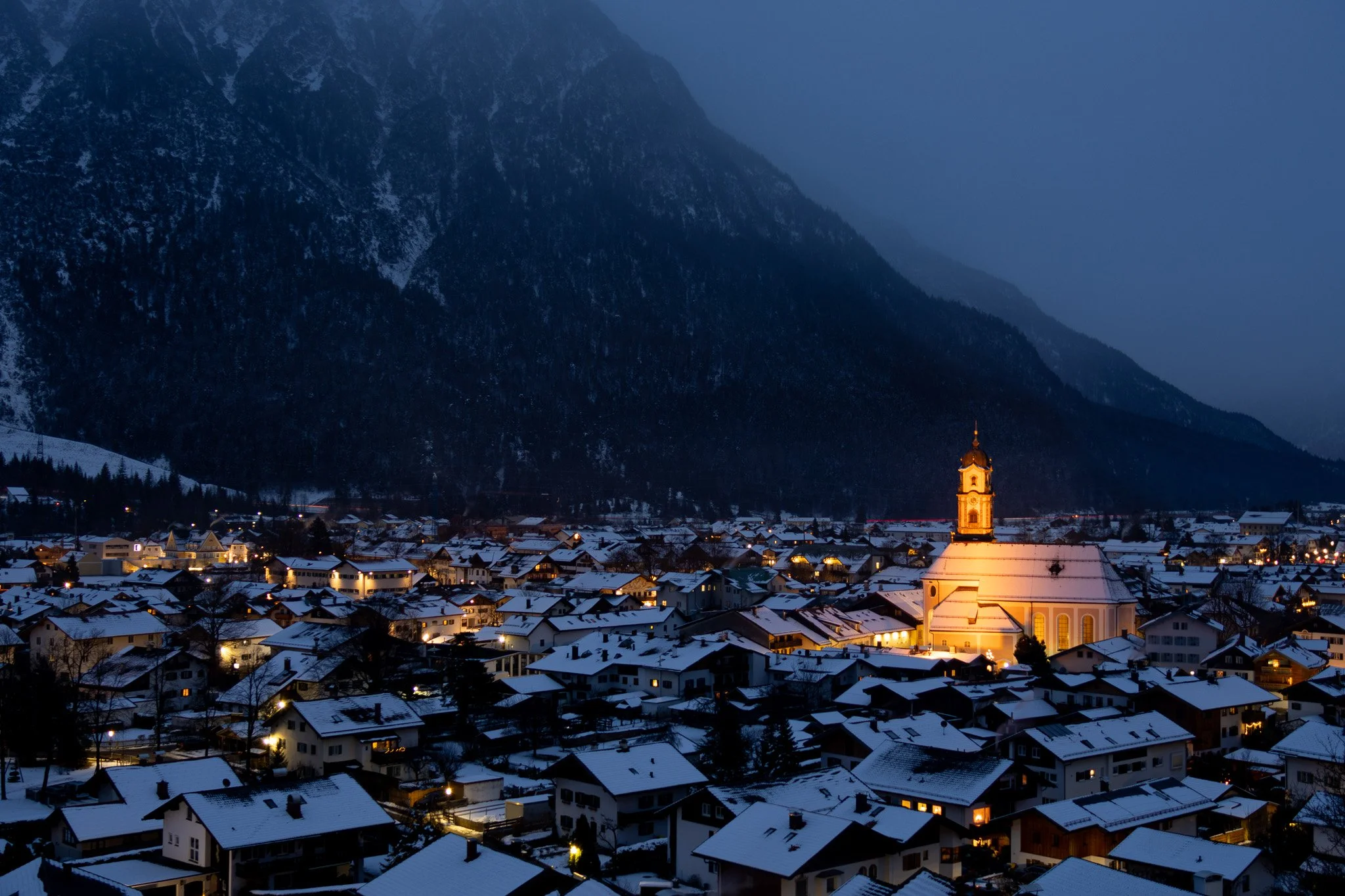 A snowy town at dusk with houses and a church illuminated, surrounded by mountains.