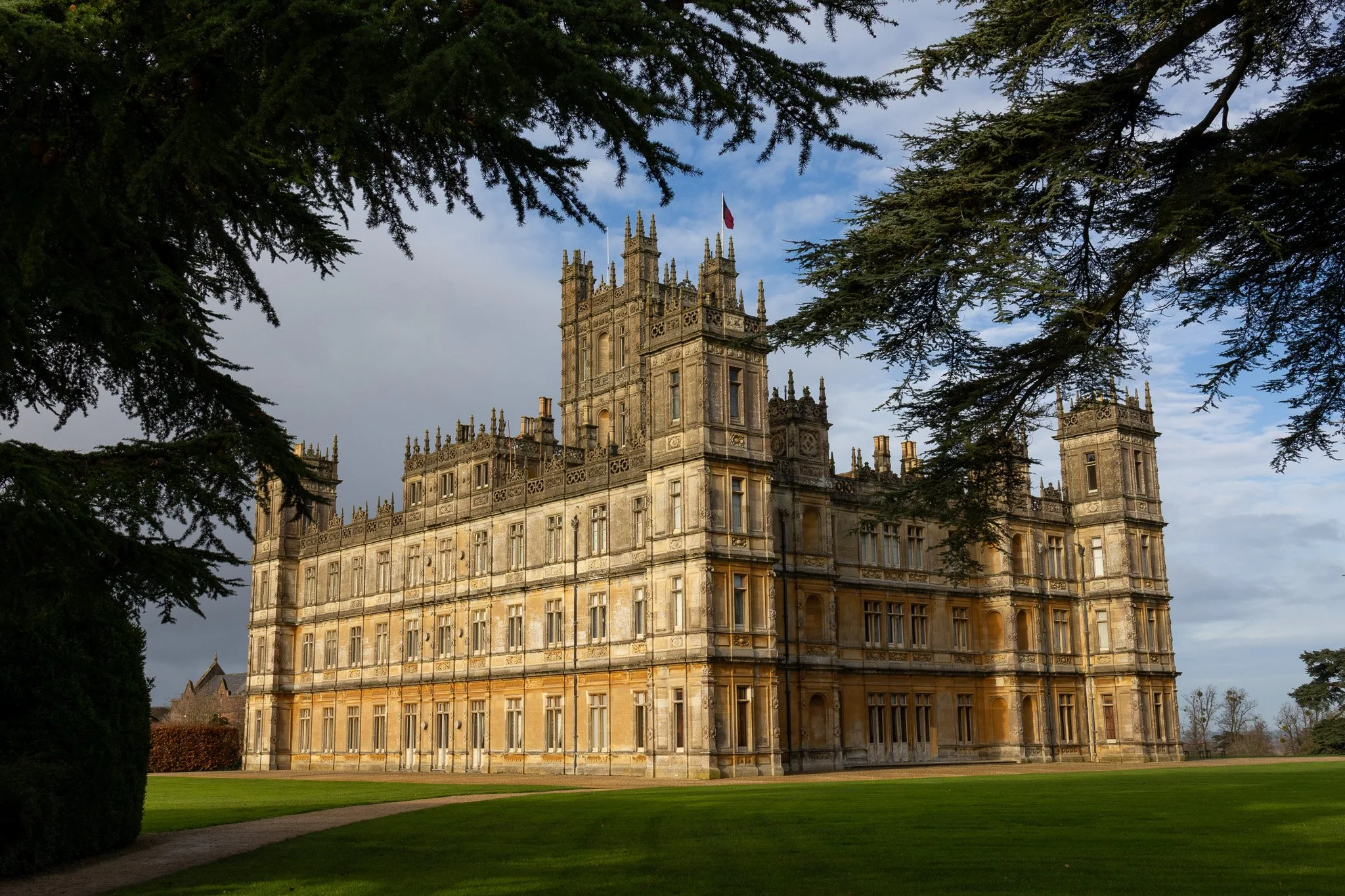 A historic castle with gothic architectural features, surrounded by greenery and framed by tree branches in the foreground, under a partly cloudy sky.