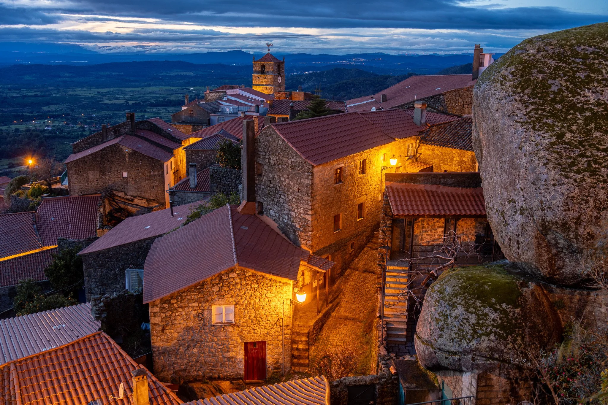 Nighttime view of a historic village with stone houses, lit by yellow streetlights, with a clock tower and a mountain range in the background.