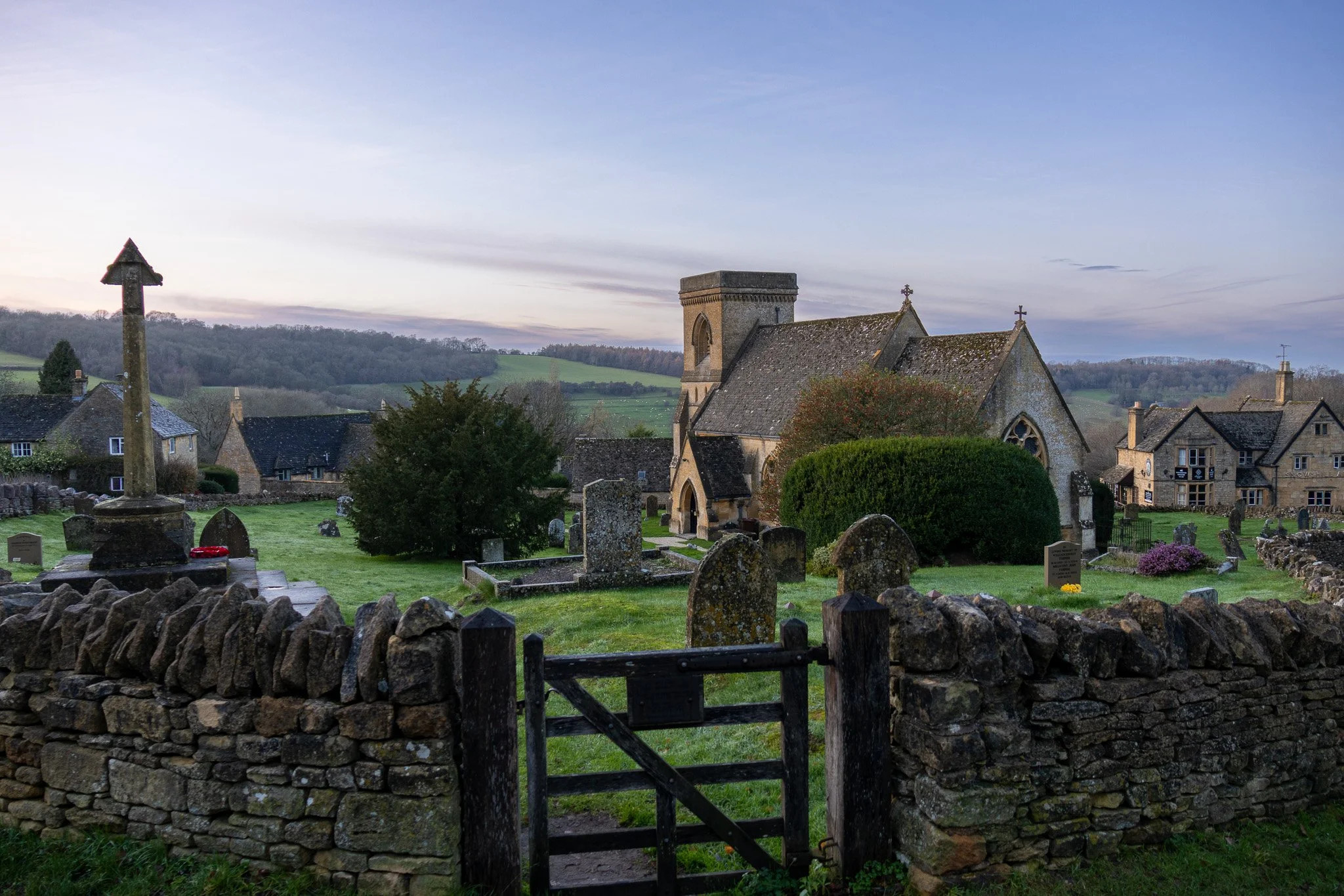 A historic church surrounded by a graveyard with headstones, a stone fence, and rolling green hills in the background during dusk.
