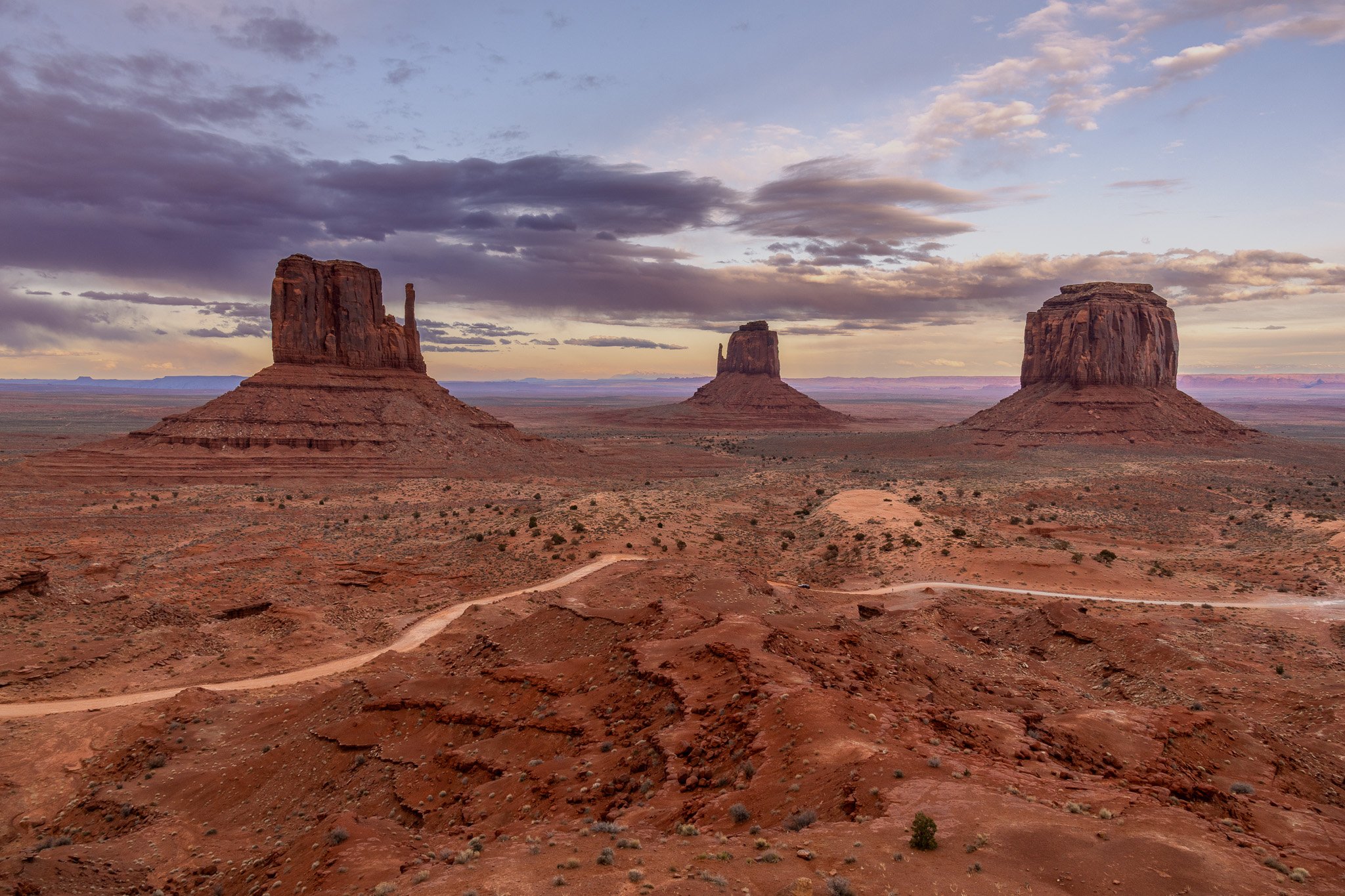 Three large sandstone buttes in a desert landscape with a cloudy sky at sunset or sunrise, with reddish terrain and sparse vegetation.