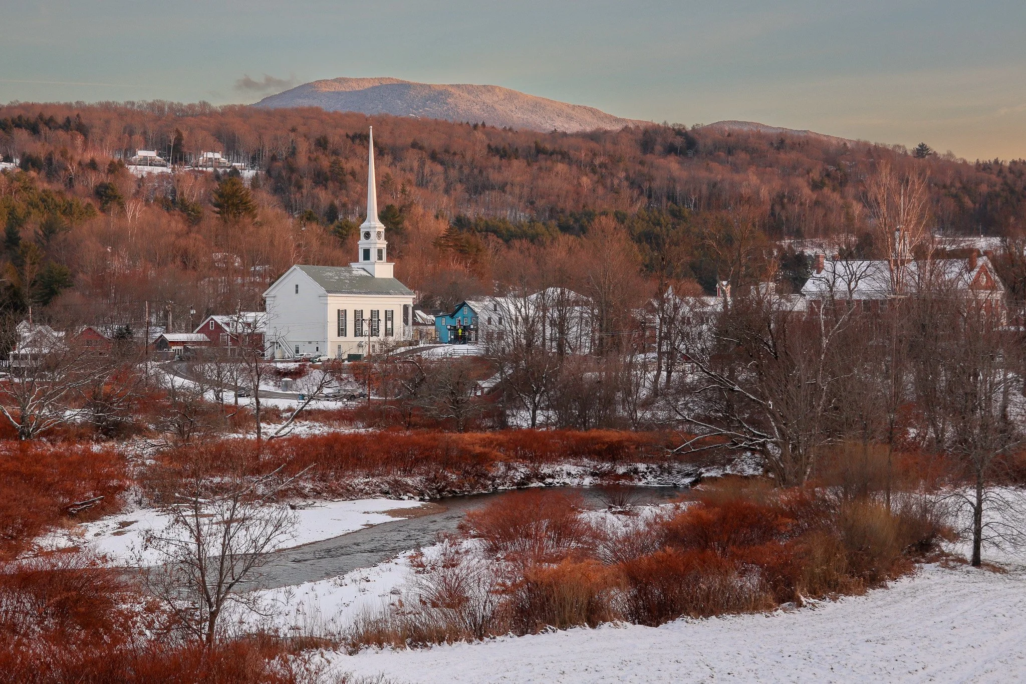 A winter landscape featuring a small town with a prominent white church with a tall steeple, surrounded by leafless trees and snow-covered ground, with mountains in the background under a clear sky.