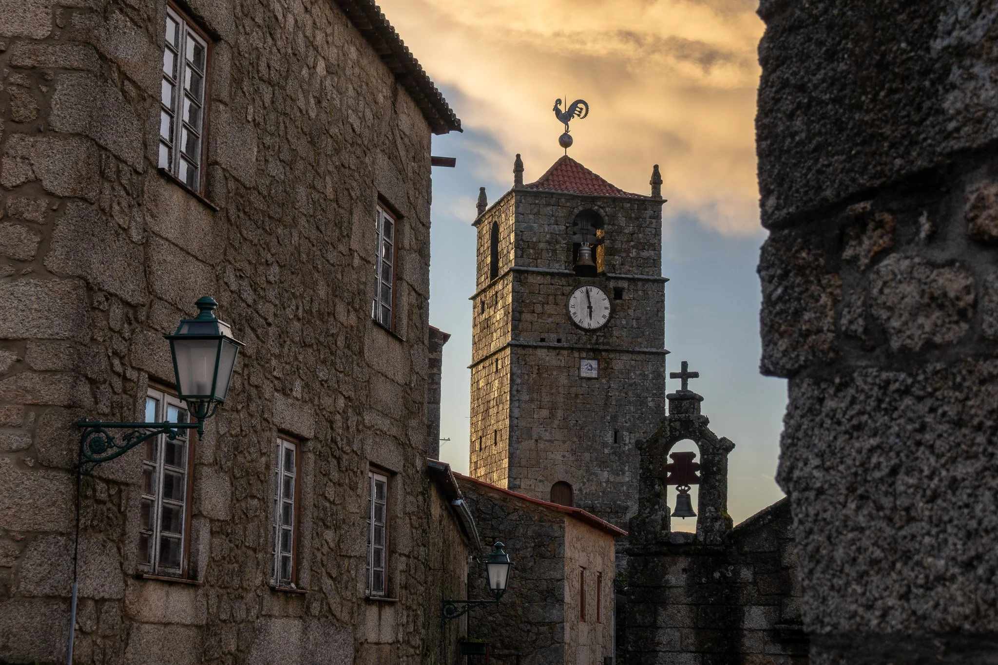 A stone clock tower with a bell and a rooster weather vane, set against a sunset sky. Surrounding stone buildings with windows and vintage street lamps.