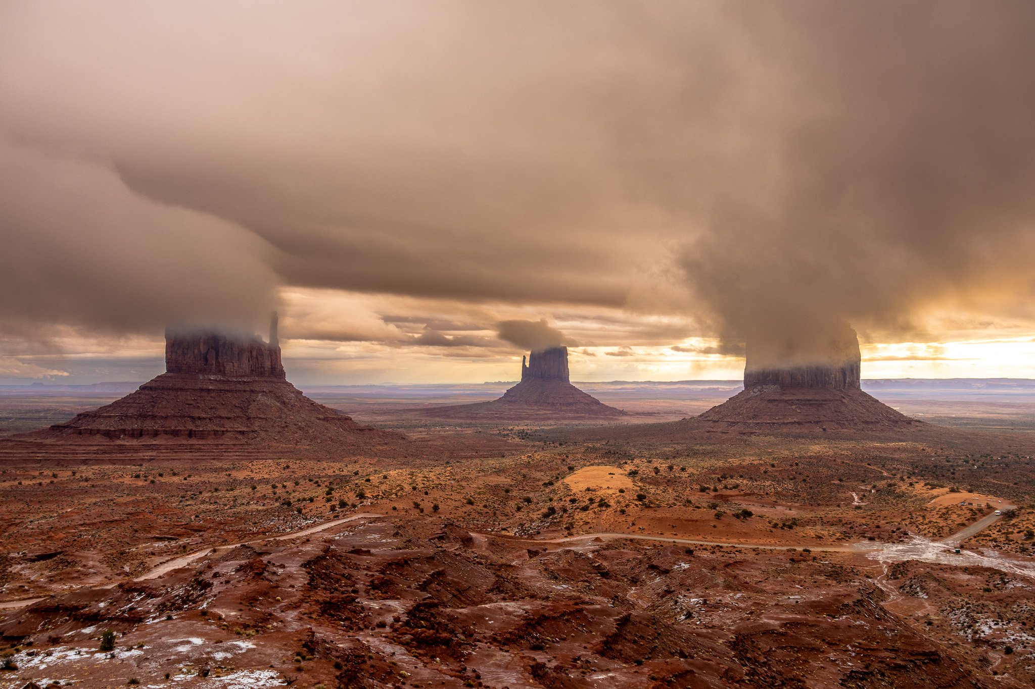 Three large sandstone buttes in Monument Valley with dark storm clouds overhead and a dirt road in the foreground