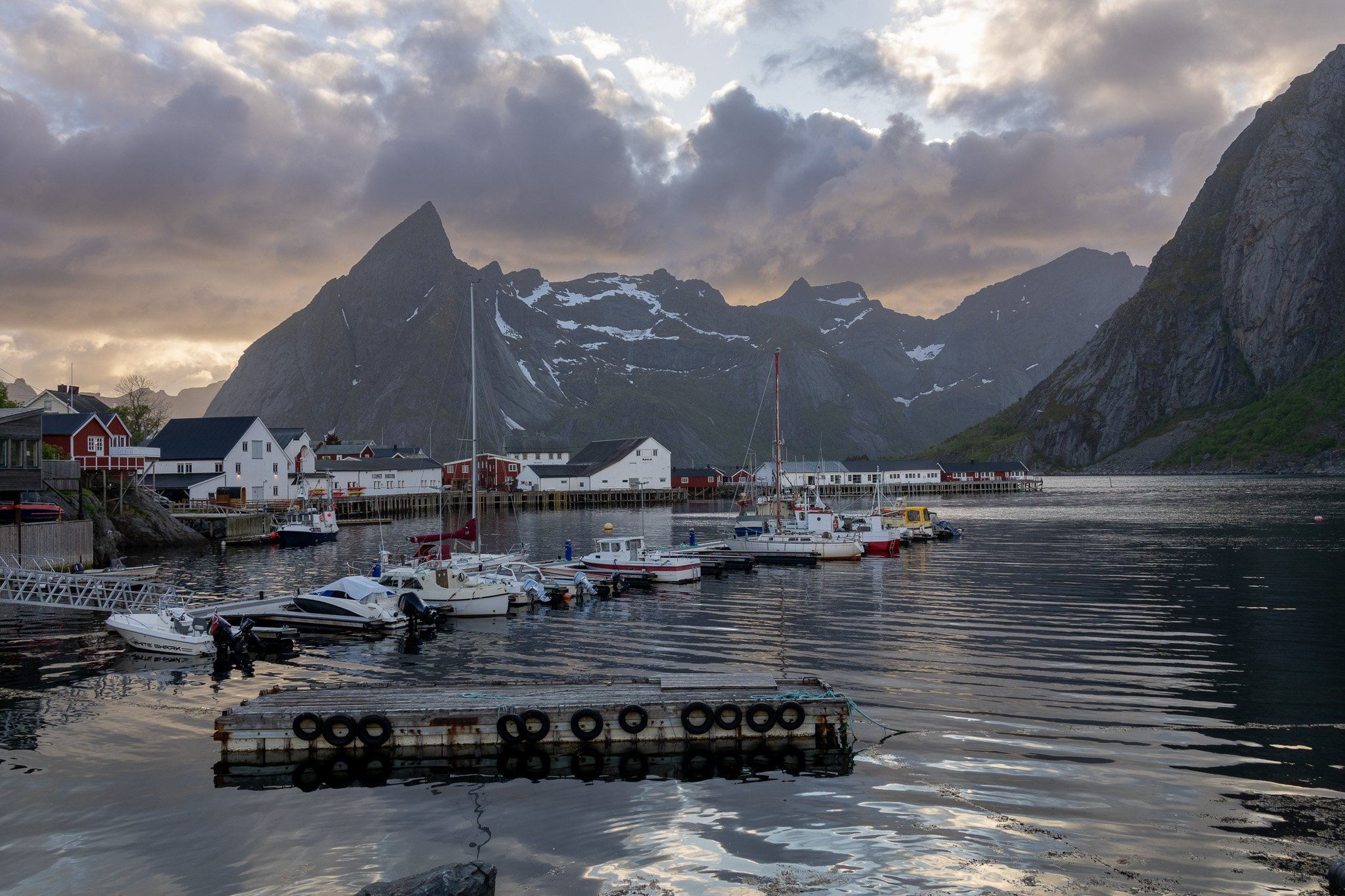 Boats docked at a marina with colorful houses under mountainous landscape during sunset.