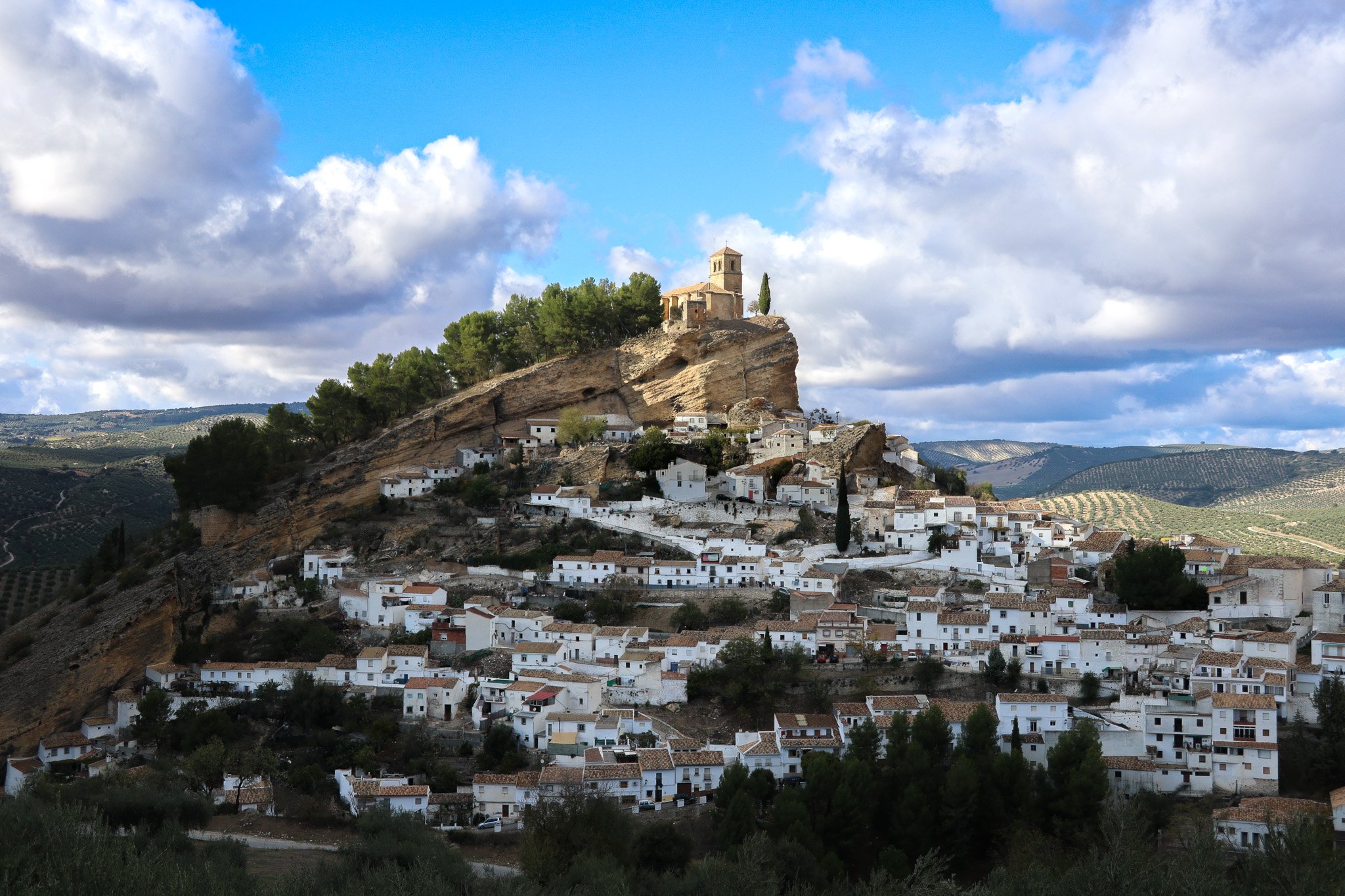 Hilly landscape with white houses and a church on top of the hill, under a partly cloudy sky.