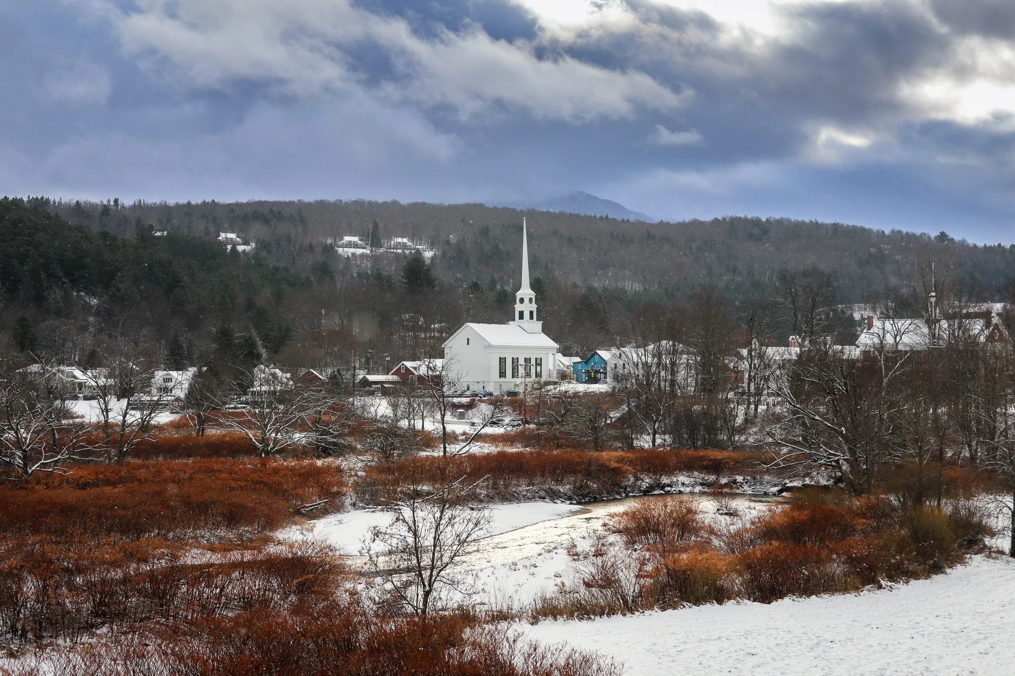A winter landscape featuring a white church with a tall steeple, surrounded by snow-covered trees and houses, with a partly cloudy sky and mountains in the background.