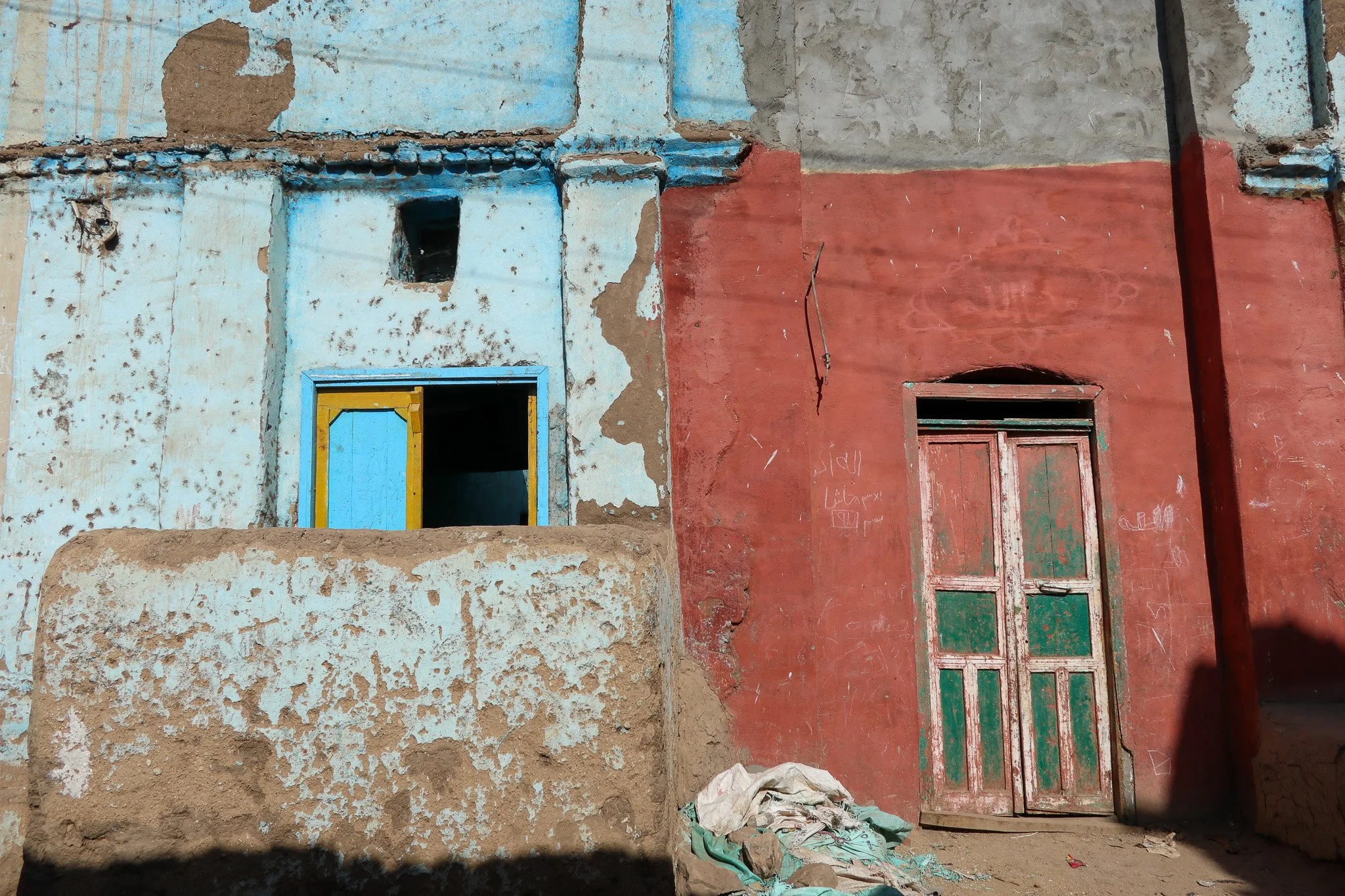 An old, weathered building with a red wall and a blue and yellow window frame. The building shows signs of wear, with patches of peeling paint and graffiti on the red wall. There is a pile of cloth on the ground and a shadow cast on the lower part of