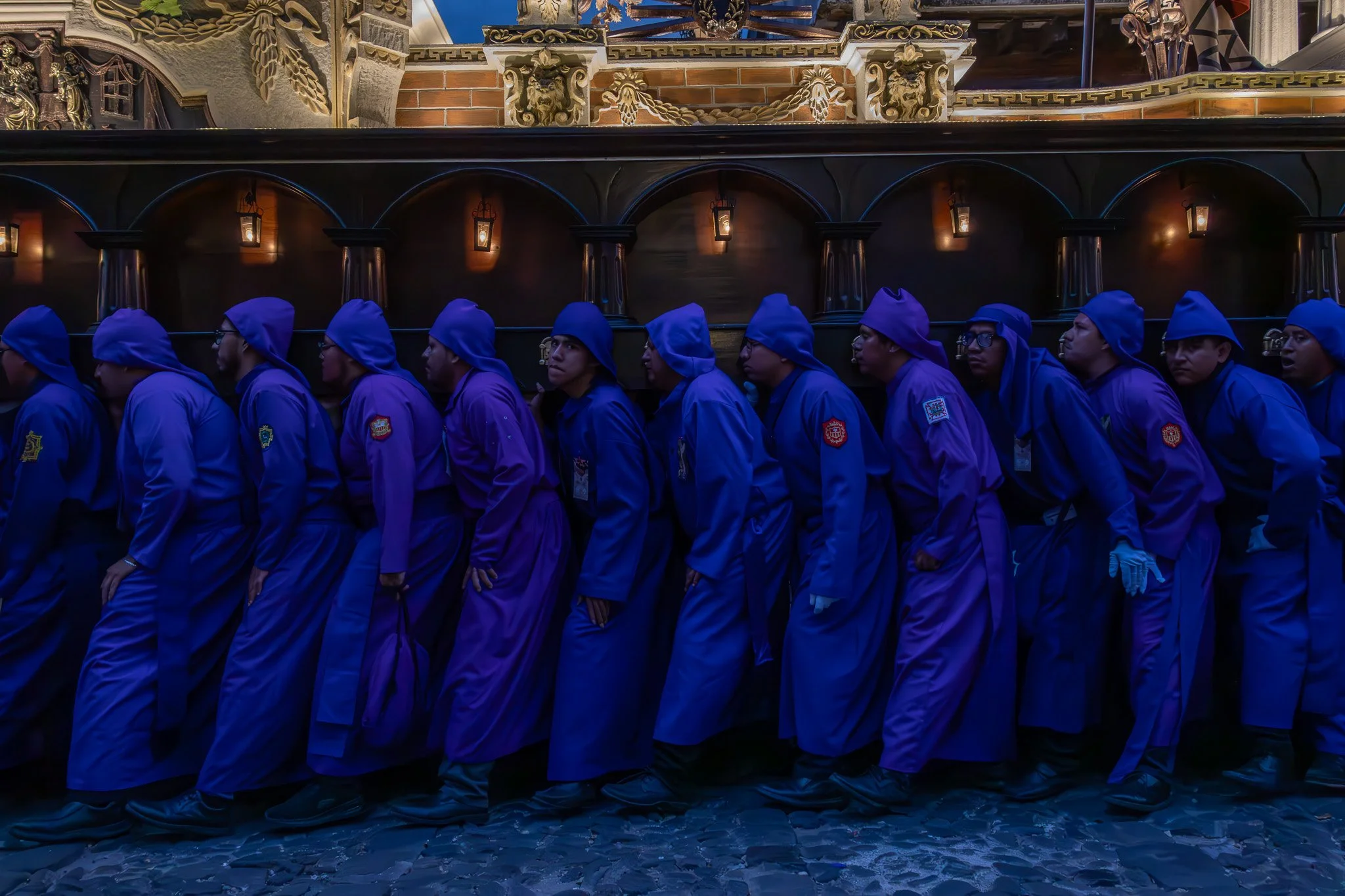 A group of people dressed in purple robes and blue hats, standing in a line inside a setting with ornate architectural details and warm lighting.