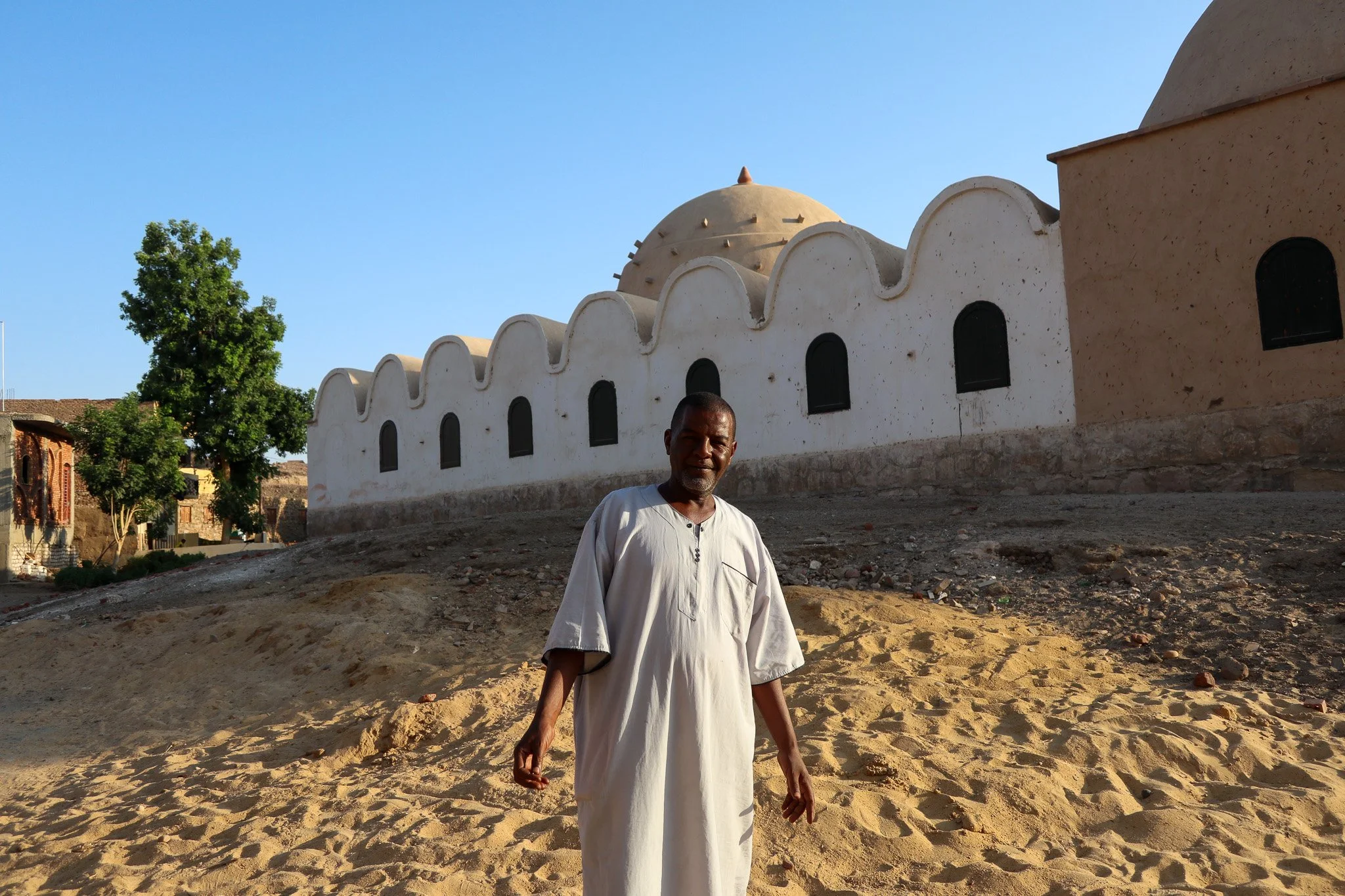 A man dressed in white traditional clothing standing on sandy ground in front of a white building with arched windows and a dome, with a clear blue sky in the background.