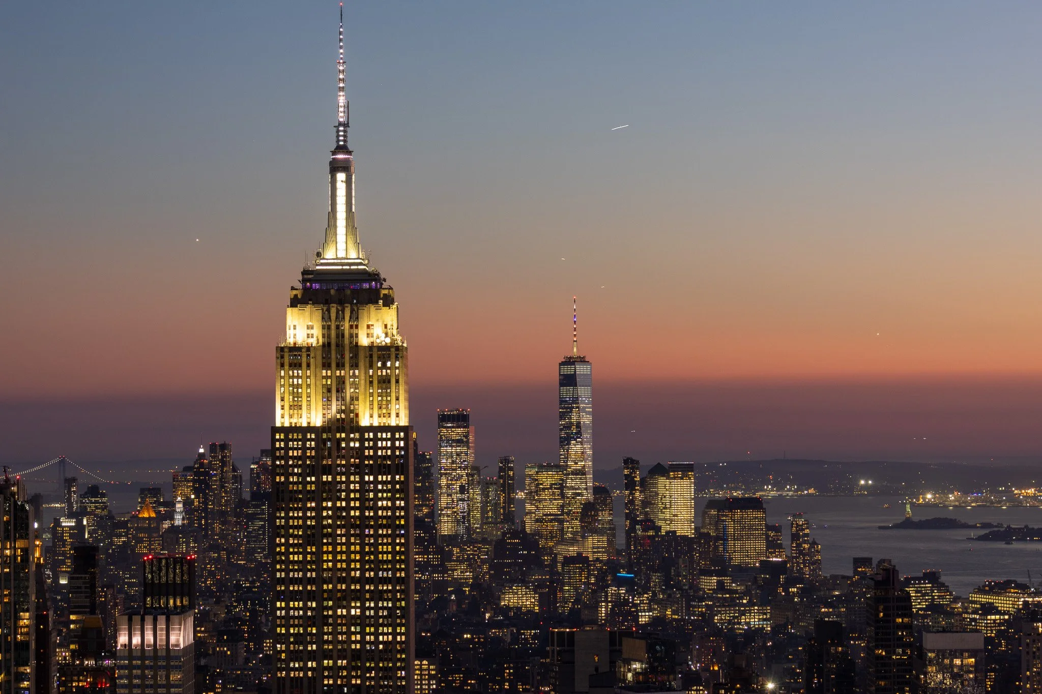 Nighttime city skyline in New York City with illuminated Empire State Building and One World Trade Center, with a colorful sunset sky and water in the background.