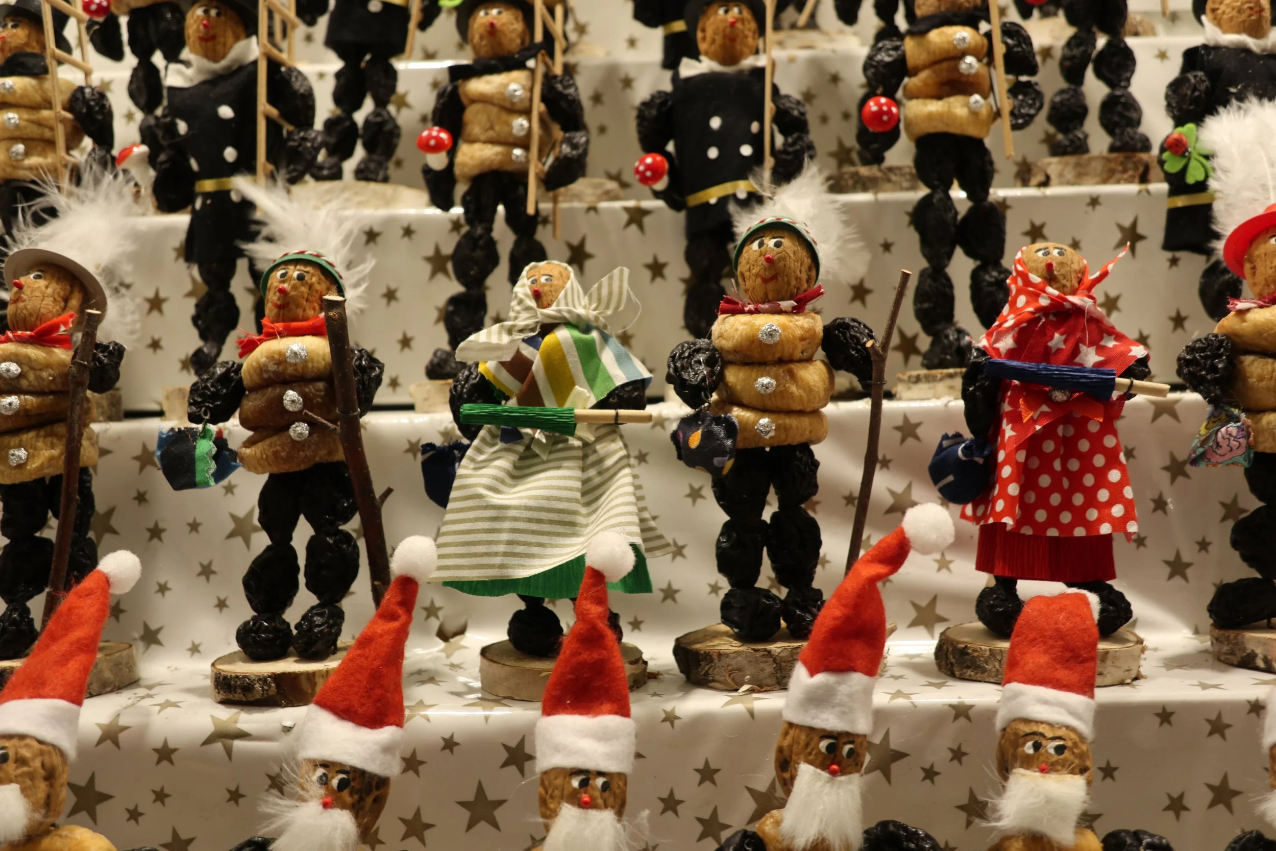 Display of Christmas figurines and decorations, including small Santa Claus figures with red hats and beards, dressed in festive clothing, and nutcracker-like figures with black hats, black and gold outfits, some holding sticks, arranged on a star-pa