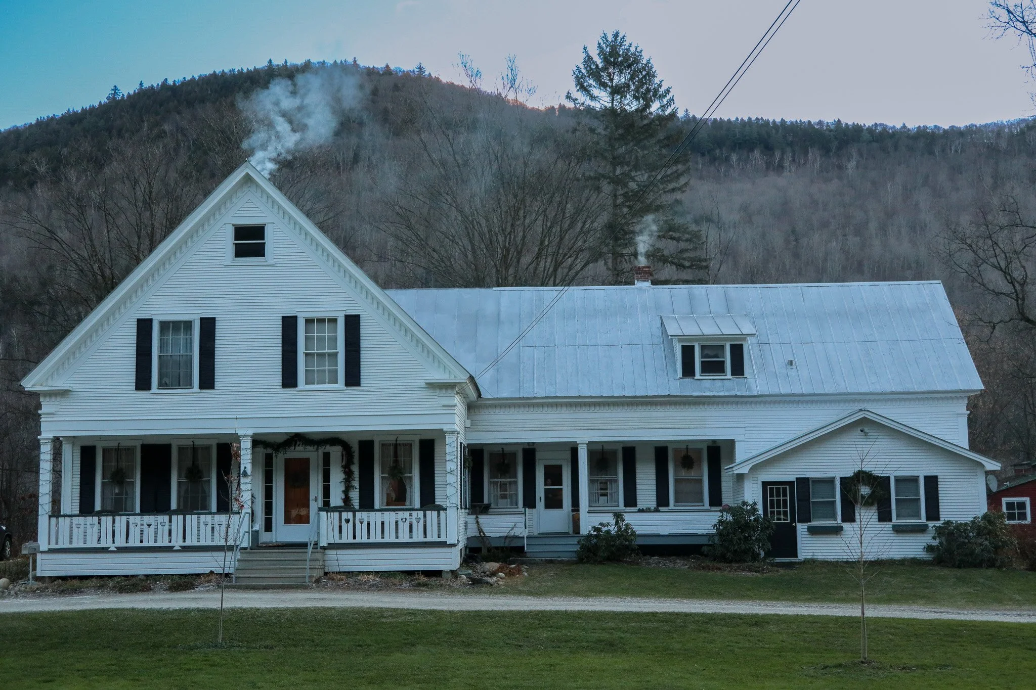 Large white house with black shutters, front porch, and metal roof, set against a wooded mountain background, with leafless young trees in the yard.