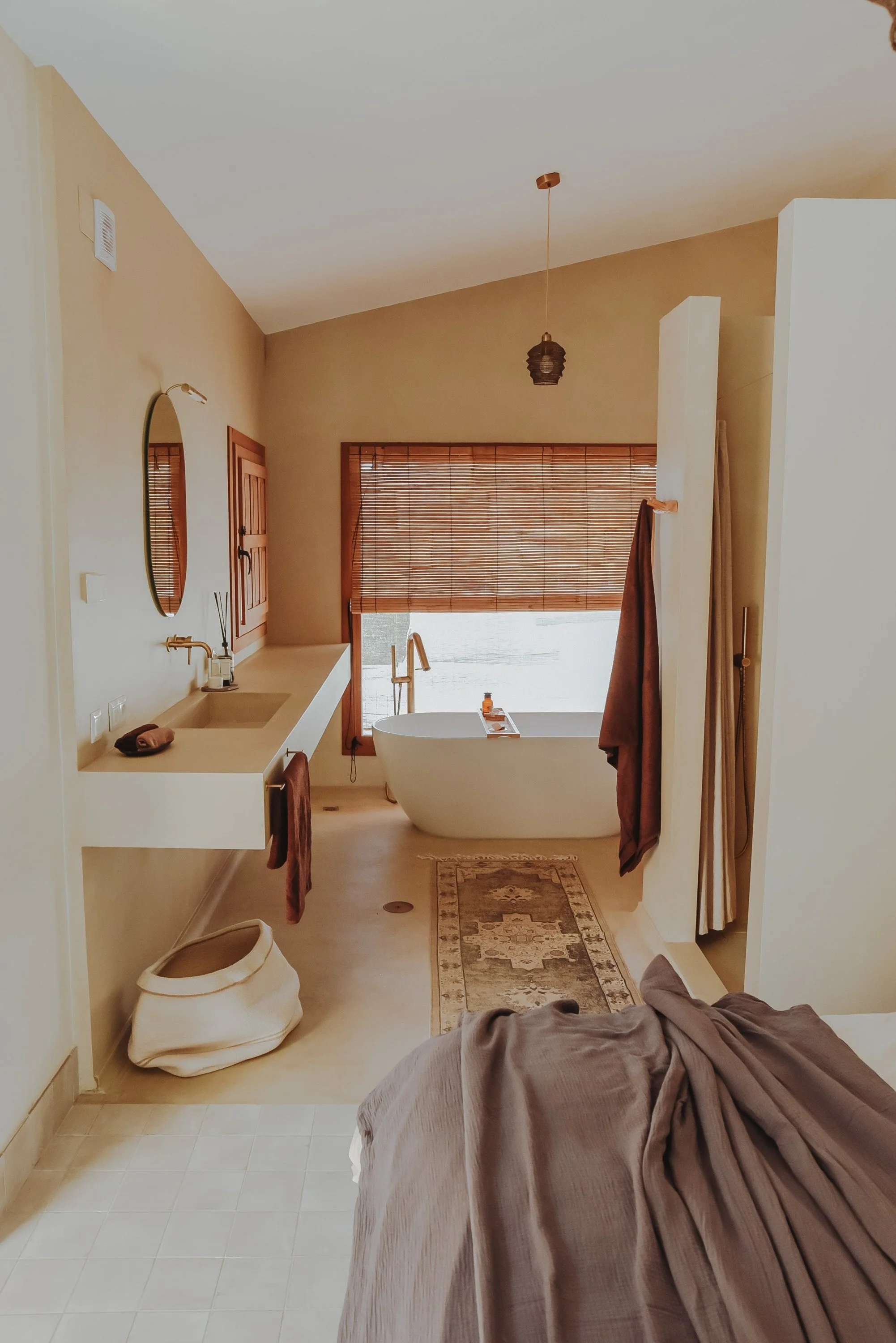 A bathroom with a bathtub in front of a large window with wooden blinds, a beige wall, and a ceiling with a hanging light. There is a sink with a mirror on the left, and towels hanging on hooks. The floor has a rug, and a brown towel or robe is draped over a surface on the right, partially covering a large white vase on the floor.