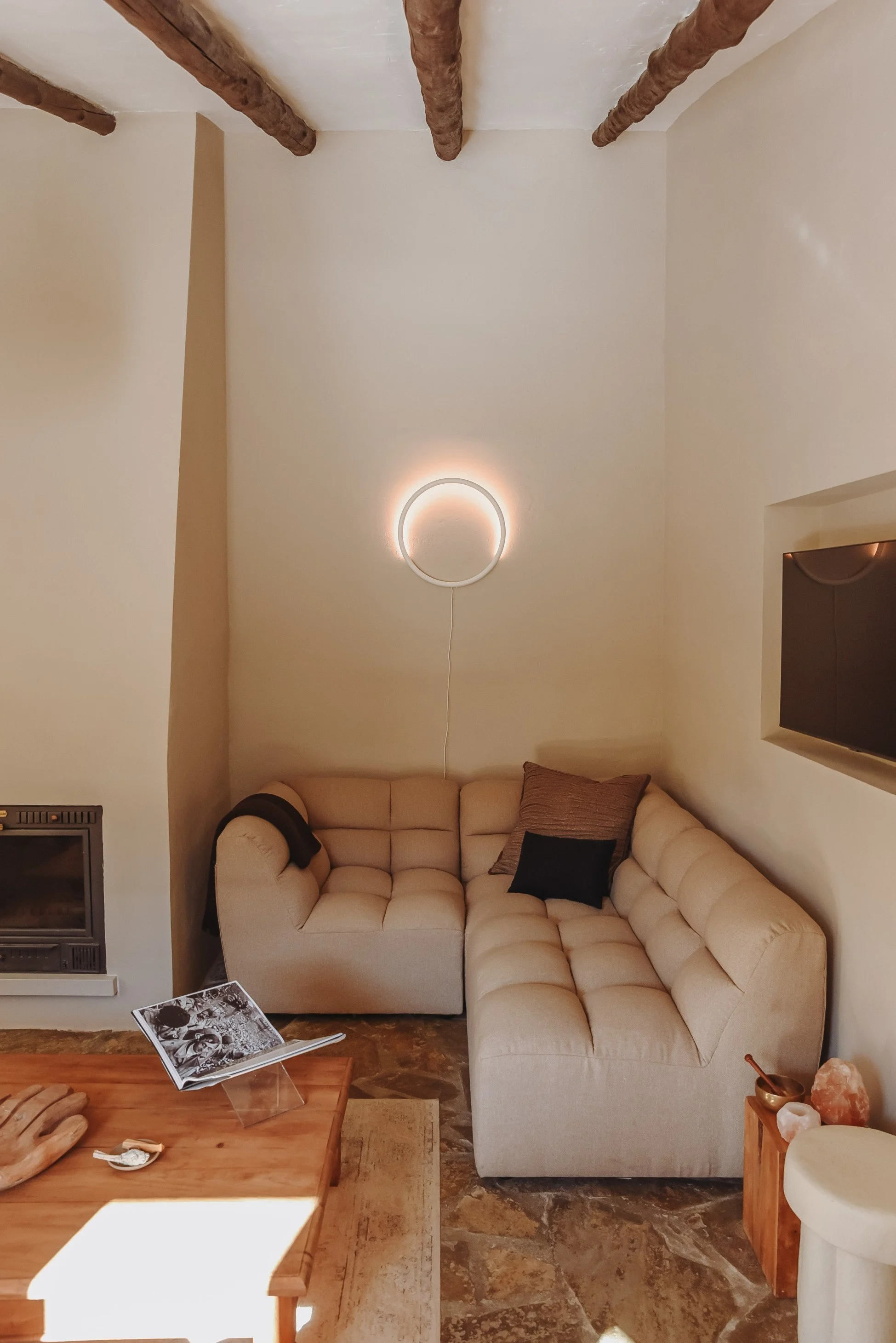 Living room with beige sectional sofa, wooden coffee table, wall-mounted TV, and copper salt lamp on side table, illuminated by circular wall light; beige walls and stone floor.