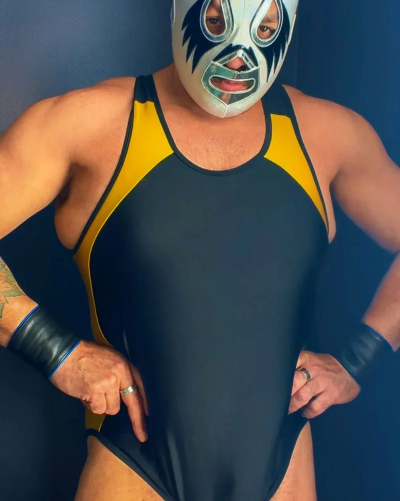 Masked luchador poses in a powerful stance with both hands on his hips. He wears a metallic mask and black and yellow leotard.
