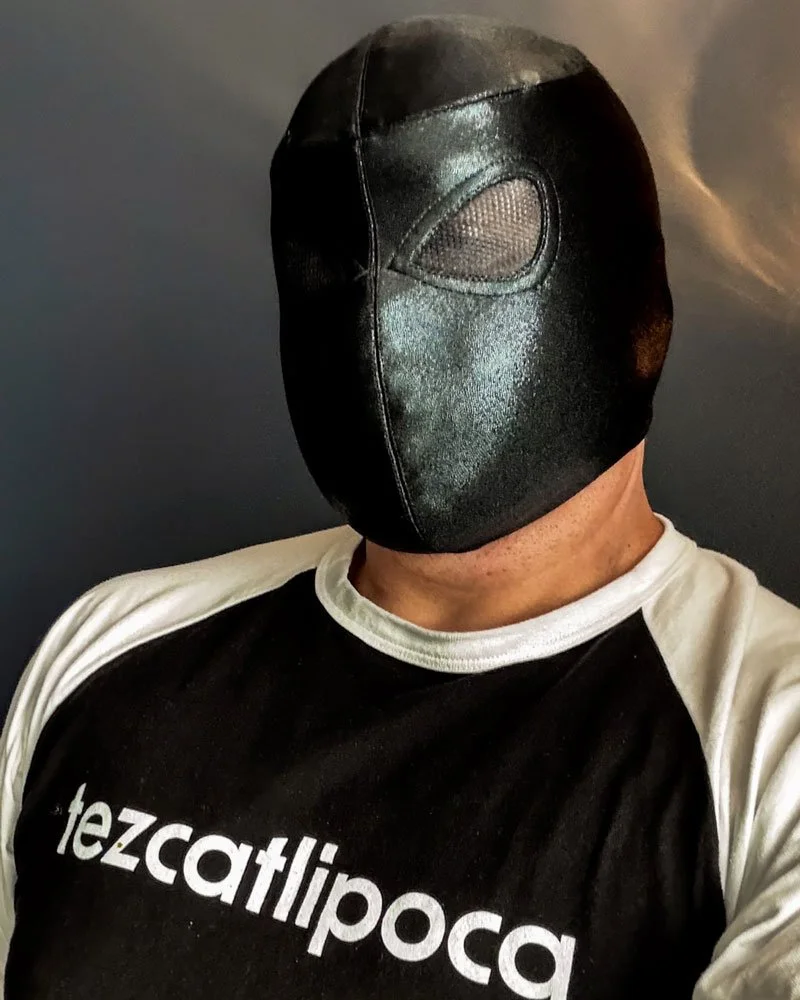 Nagual the masked luchador poses in a black mask and black and white t-shirt that reads "Tezcatlipoca"