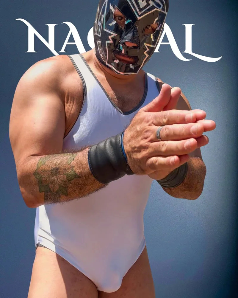 The masked luchador known as Nagual poses in a black and white mask and white leotard as he clasps his hands. He stands before a gray background and behind him the word "Nagual" can be partially seen.