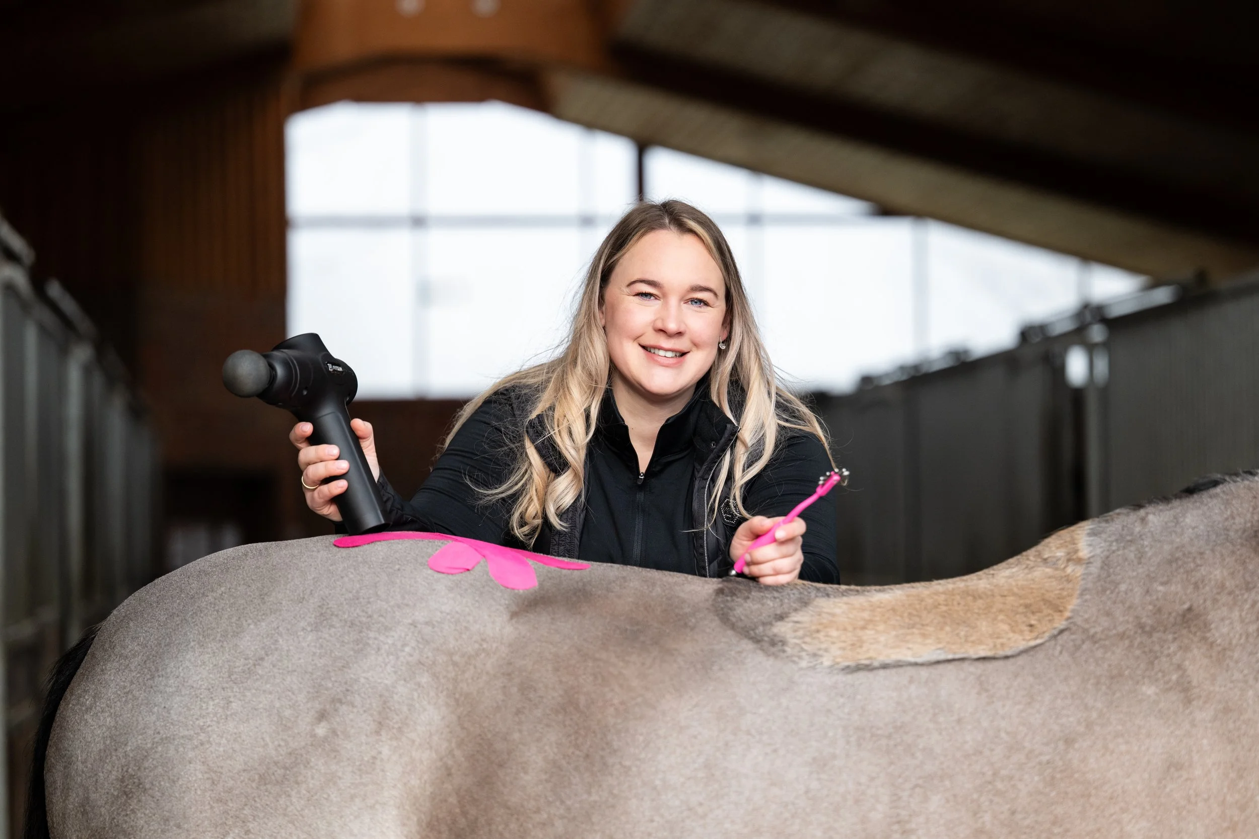 Eine blonde Frau mit einem schwarzen Pullover streichelt ein braunes Pferd im Stall, während sie eine Massagegerät in der Hand hält. Das Pferd hat eine rosa Blume auf dem Rücken.