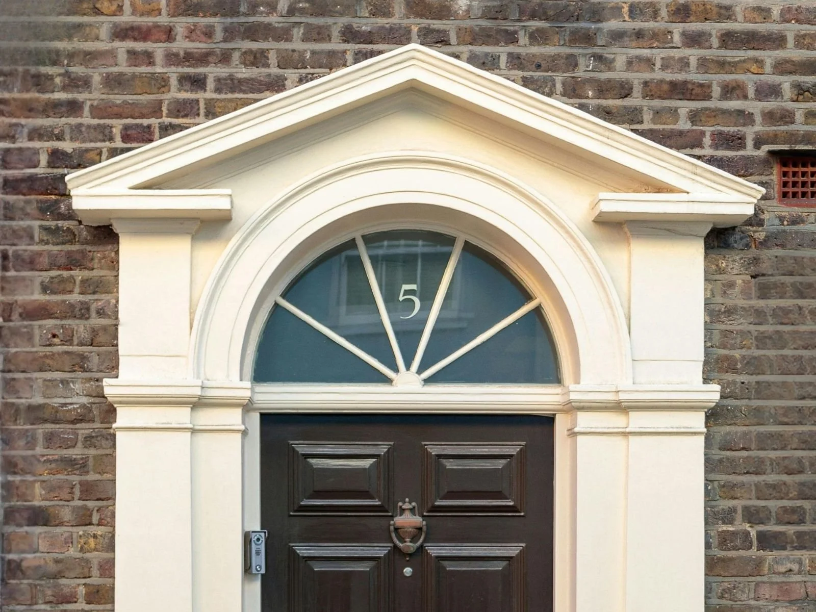 A dark wooden front door with decorative panels and a door knocker, set in a brick wall with an arched window and white architectural trim above.