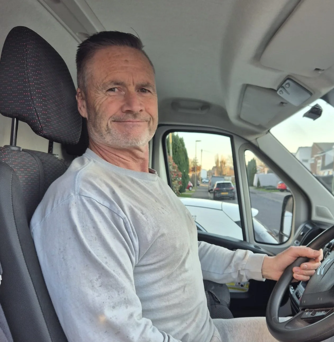 A man with gray hair and beard smiling while sitting in the driver's seat of a van, holding the steering wheel, with a suburban neighborhood visible through the window.