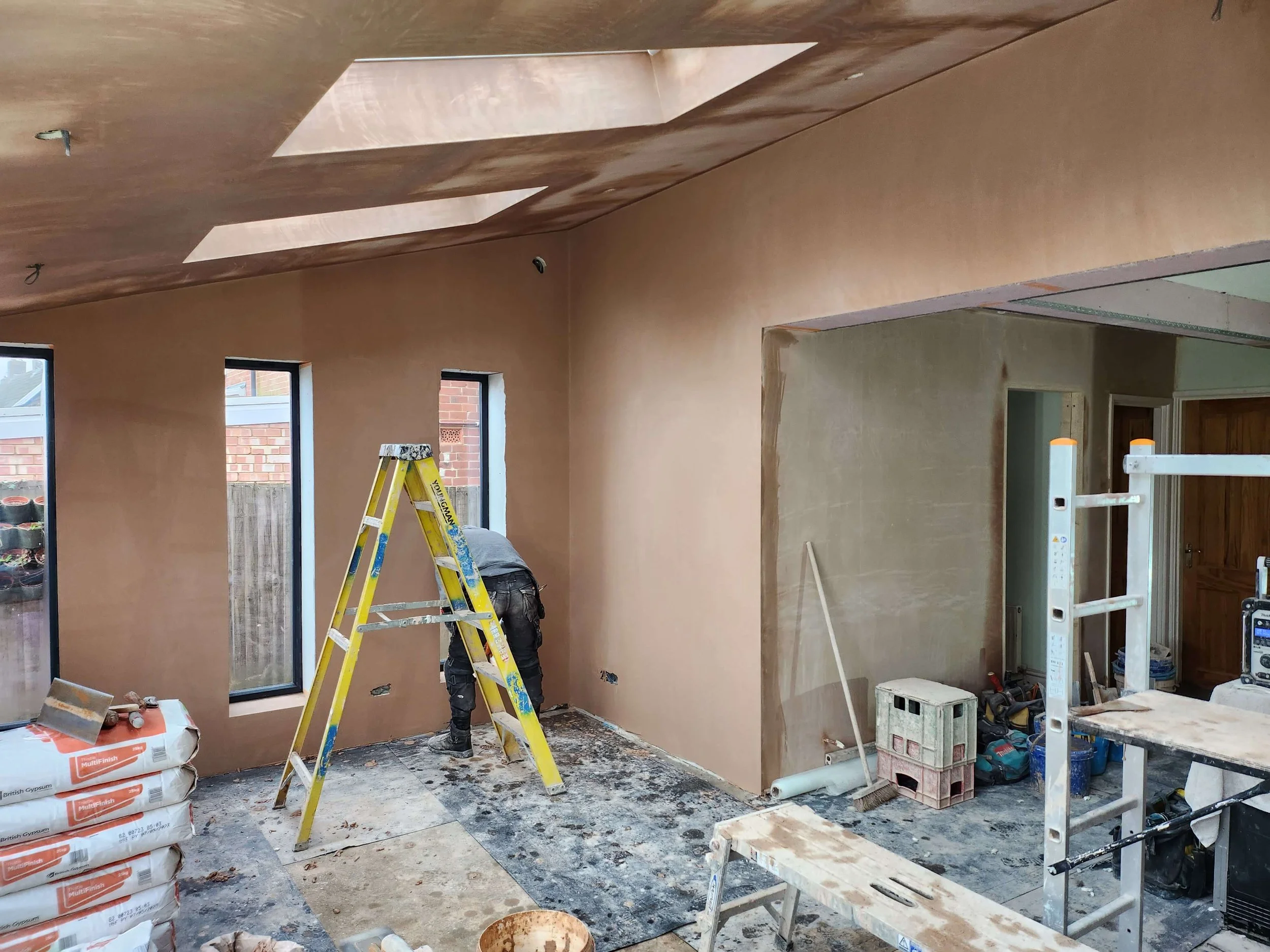 Interior of a house under construction with drywall being installed on the ceiling and walls, a ladder, construction tools, and materials such as bags of drywall compound and various supplies visible.