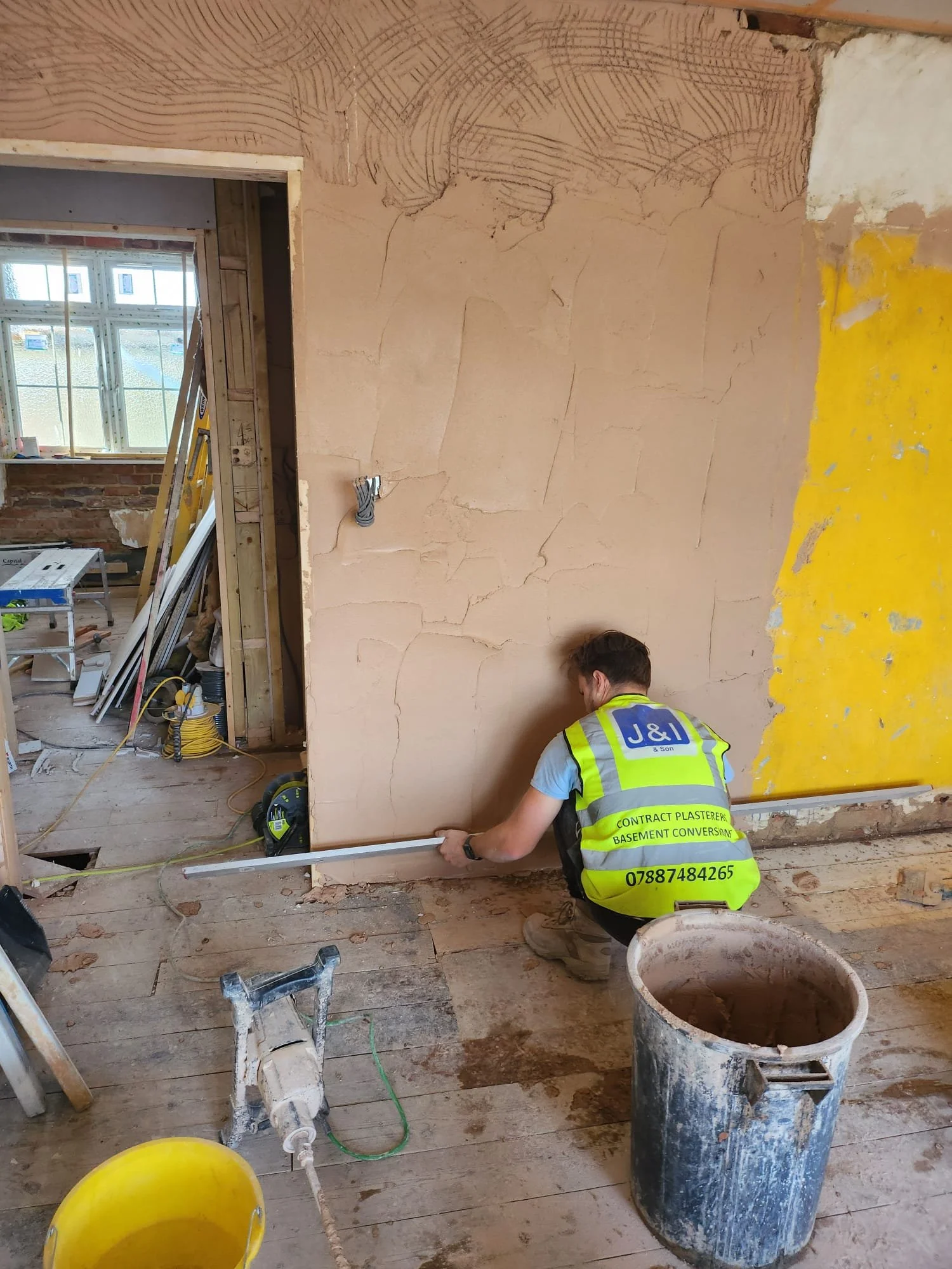 A worker in a yellow safety vest measures a large piece of drywall on a construction site. The room has partially finished walls, exposed wooden floor, and construction tools and materials scattered around. A window in the background lets in natural light.
