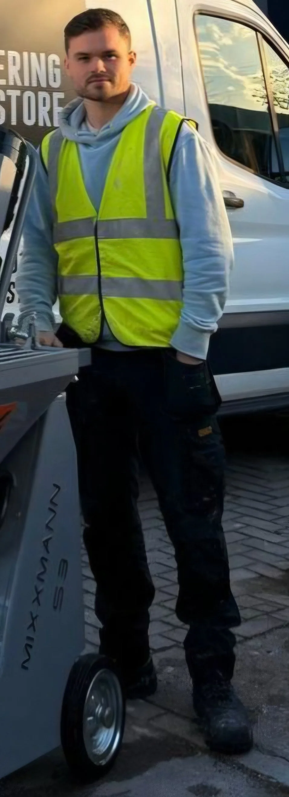 A young man wearing a yellow reflective safety vest and black work pants standing outdoors beside a mobile food cart with a Nespresso logo, with a white vehicle and a building in the background.