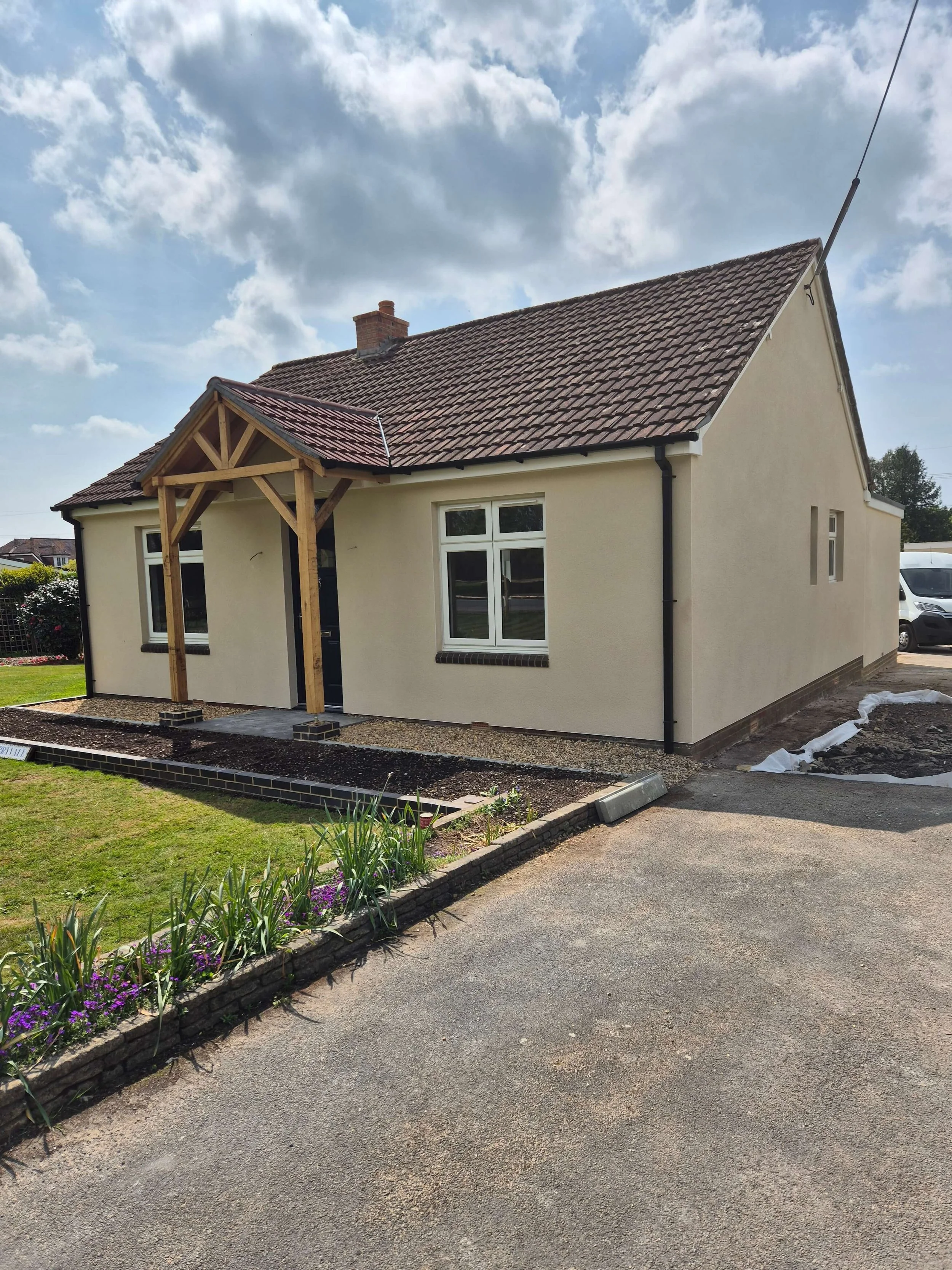 New house with beige exterior walls, dark brown roof, window, new front porch with wood frame, and a small garden.