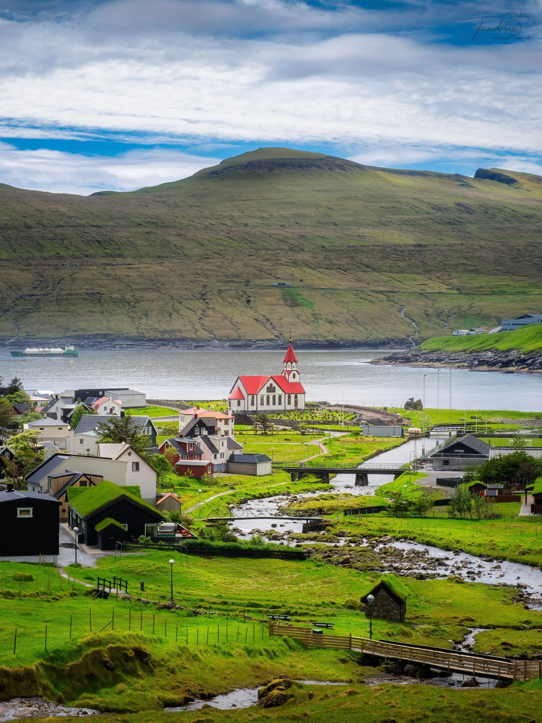 Sandav&aacute;gur p&aring; &oslash;en V&aacute;gar. Den hvidmalede kirke med det r&oslash;de tag har st&aring;et her siden 1917 &mdash; og regnes som en af de smukkeste p&aring; F&aelig;r&oslash;erne. Med gr&aelig;st&oslash;rvstagene, fjorden i baggr