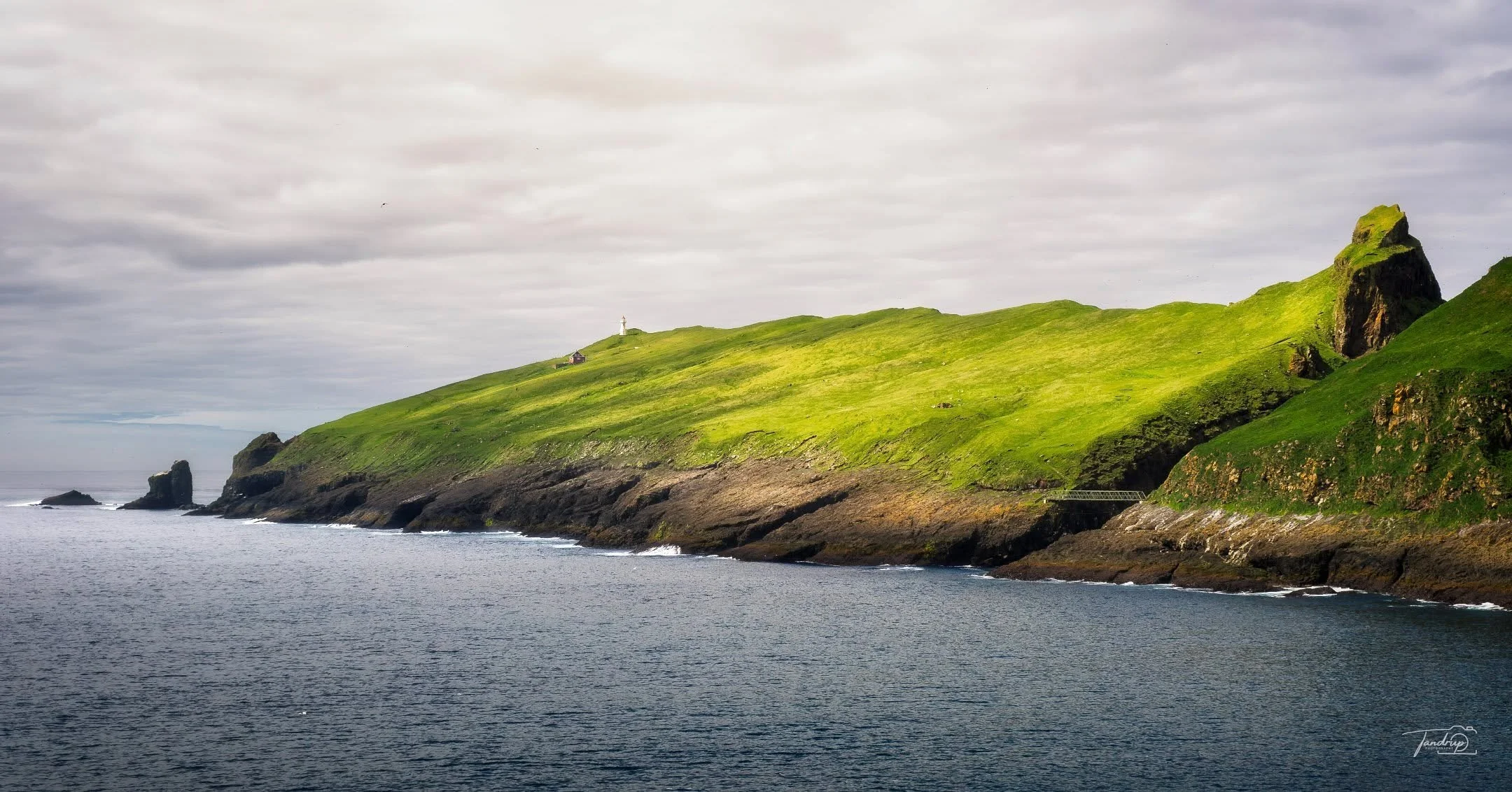 Mykines &ndash; den vestligste &oslash; p&aring; F&aelig;r&oslash;erne (og Lundeland), med Mykinesh&oacute;lmur og fyrt&aring;rnet og fyrmesterboligen fra 1909 i baggrunden. Broen til h&oslash;jre forbinder de to dele over kl&oslash;ften H&oacute;lmg