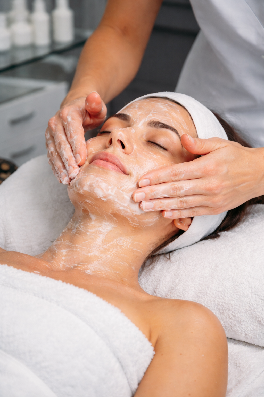 A woman receives a facial treatment at a spa, with a cream or mask being applied to her face by a professional.