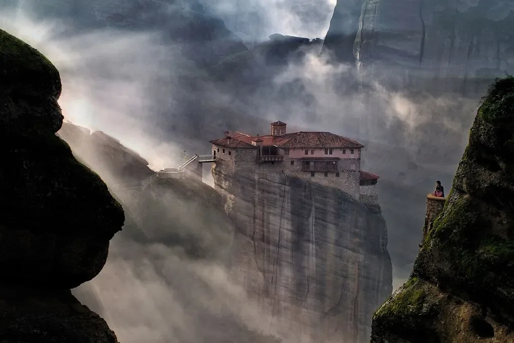 A monastery in a remote mountain region of the world with a monk looking at it from afar as the clouds dance around it and the sunlight streams through them