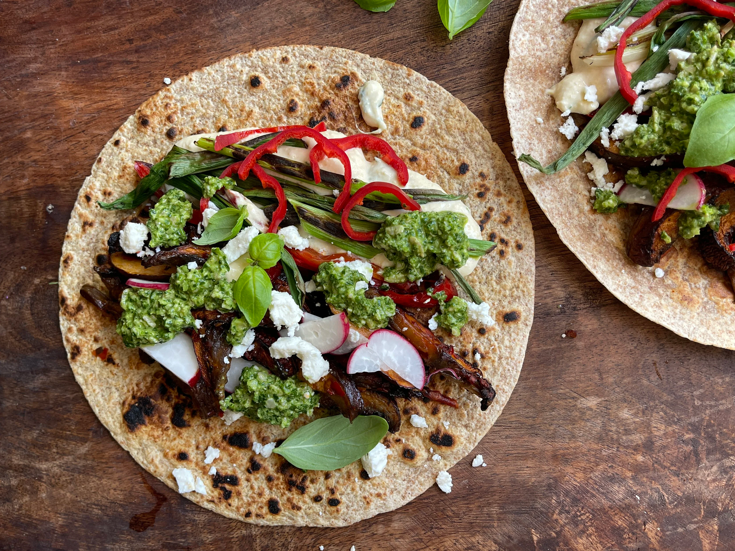 Two mushroom shawarma wraps on a wooden board topped with caramelized portobellos, bright green pistachio pesto, charred scallions, feta, and sliced radishes, inspired by the novel Circe