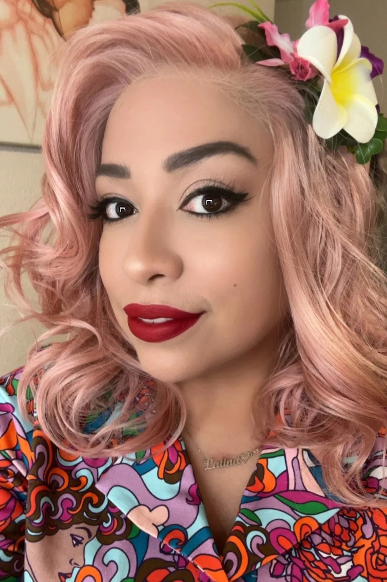A woman with pink, curly hair wearing bright makeup with red lipstick, and a colorful patterned top. She has a flower hair accessory and a necklace that says 'LATINA'.