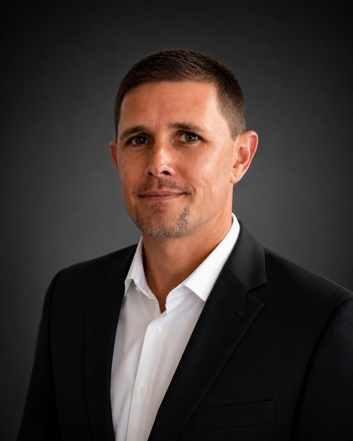 Professional headshot of a man with short brown hair, wearing a black suit and white dress shirt, against a dark gray background.