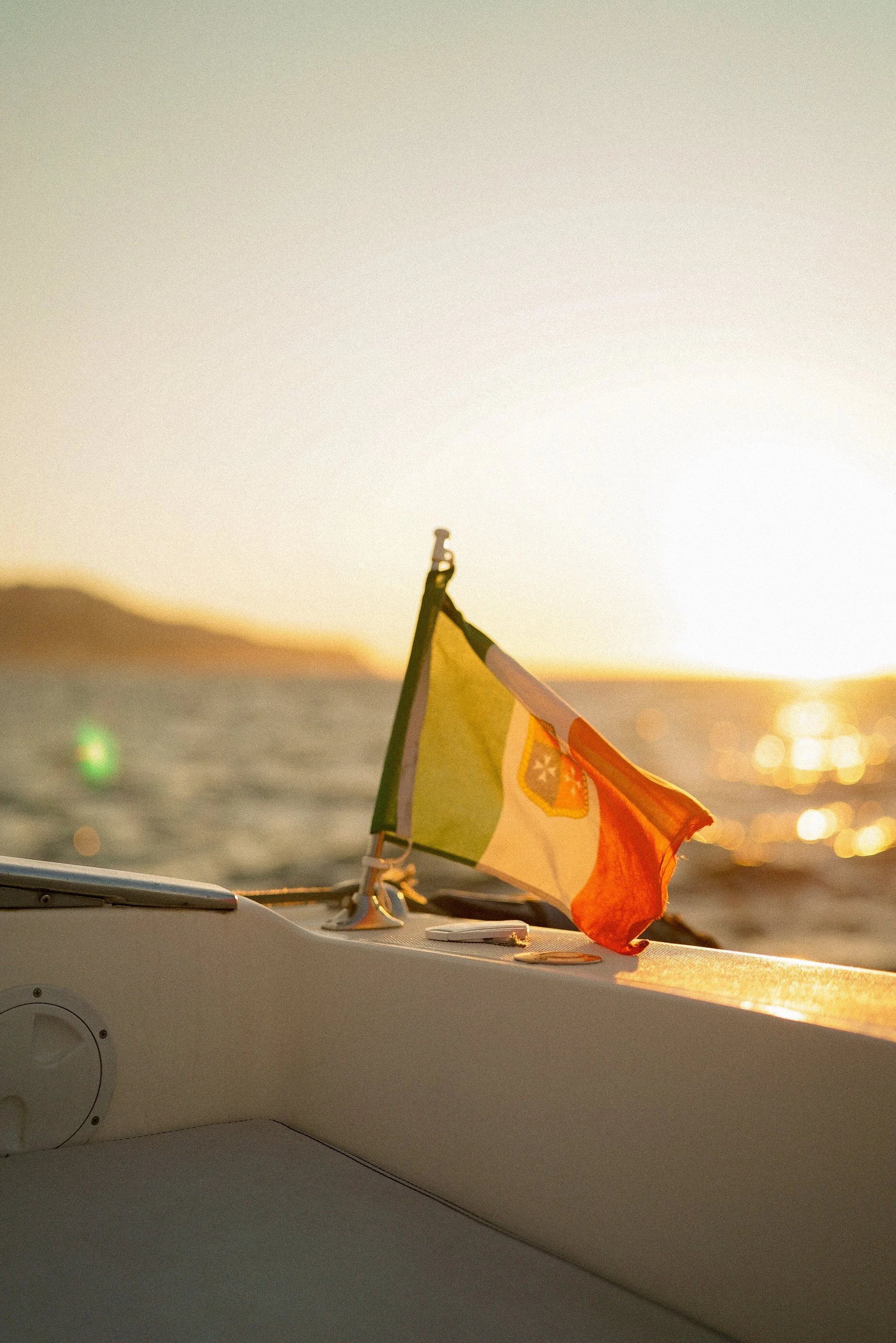 A small orange, yellow, and green sailboat flag on a boat during sunset, with a blurred island and water in the background.