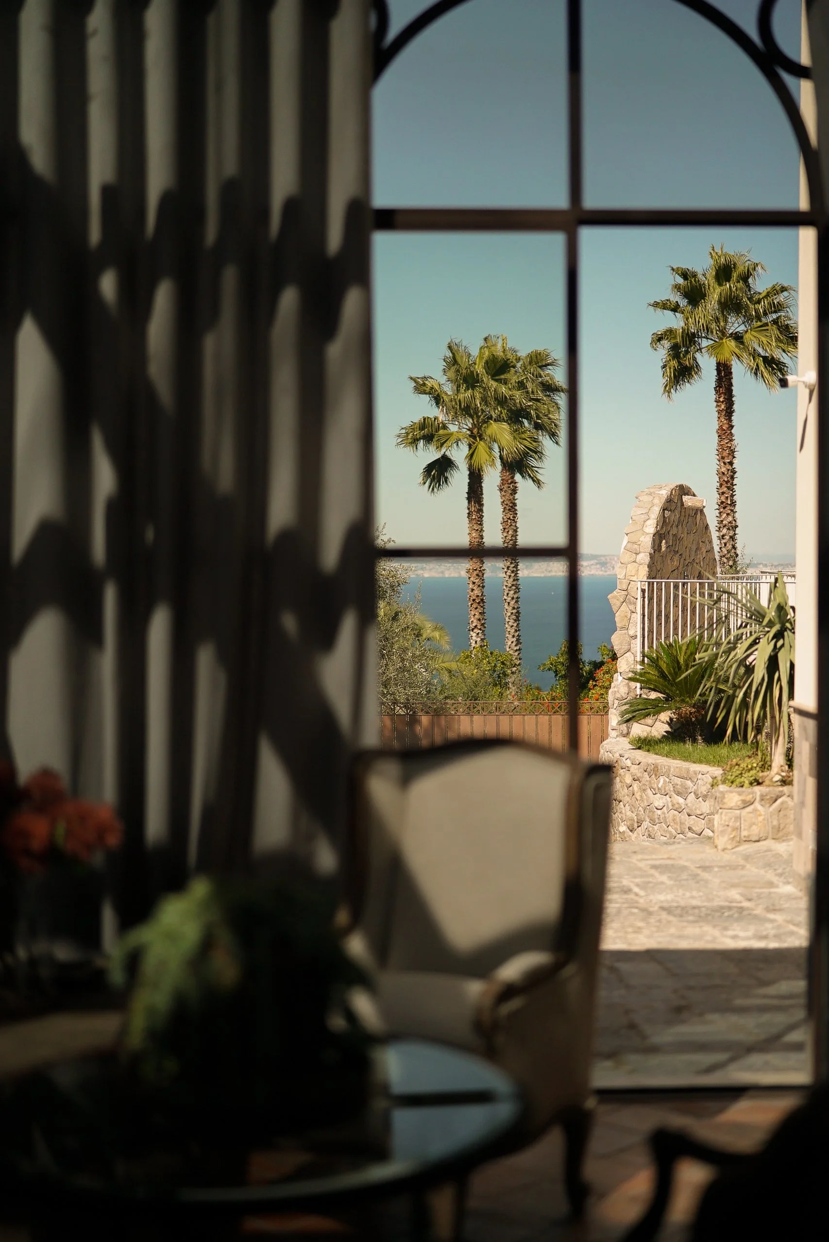 A scenic view of three palm trees seen through a window with curtain on the left and grid style window bars. The scene includes a stone structure and a fence, with water and land in the background under a clear sky.