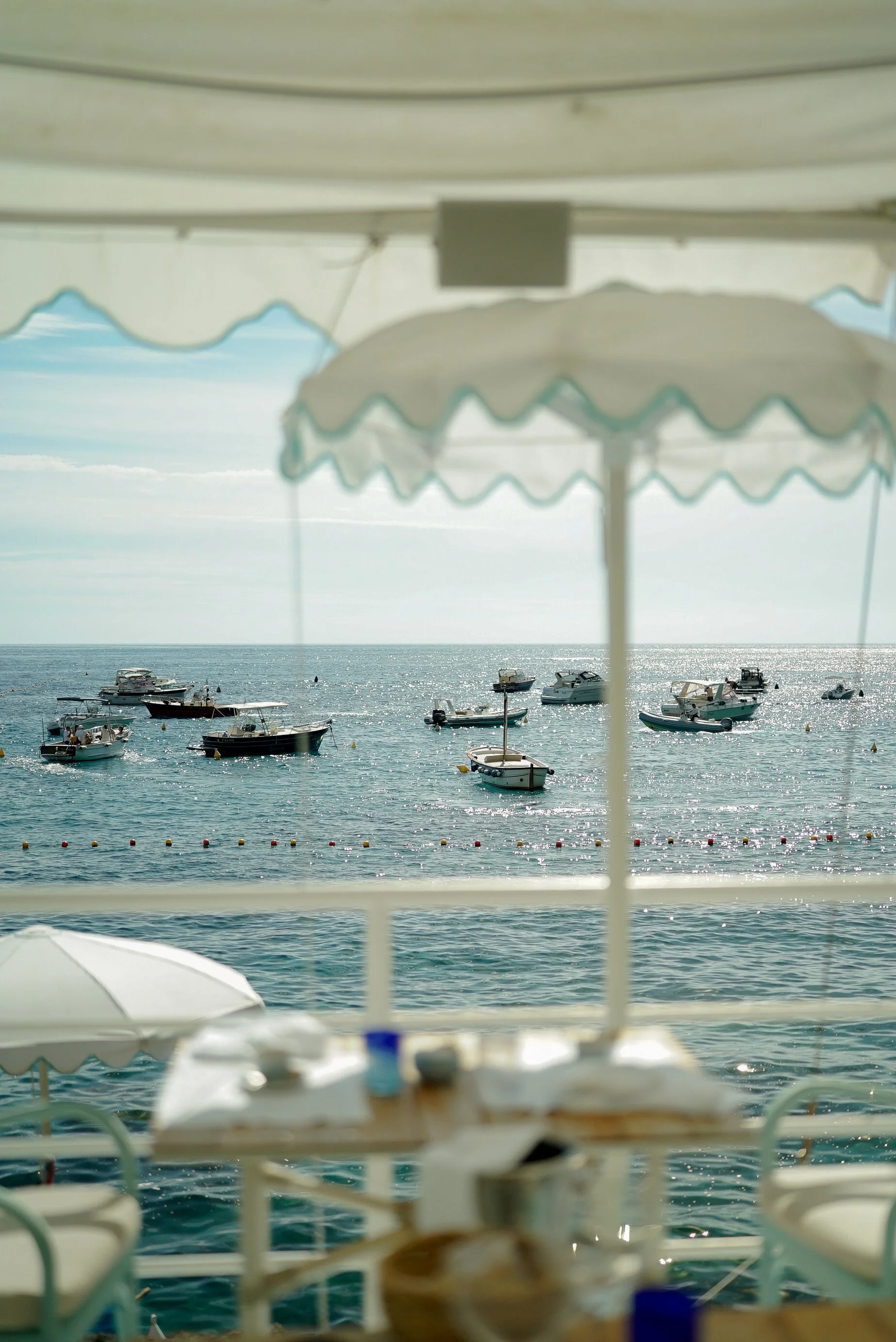 View of a seaside marina with boats anchored, seen through a frame of white and blue striped umbrellas on a terrace.