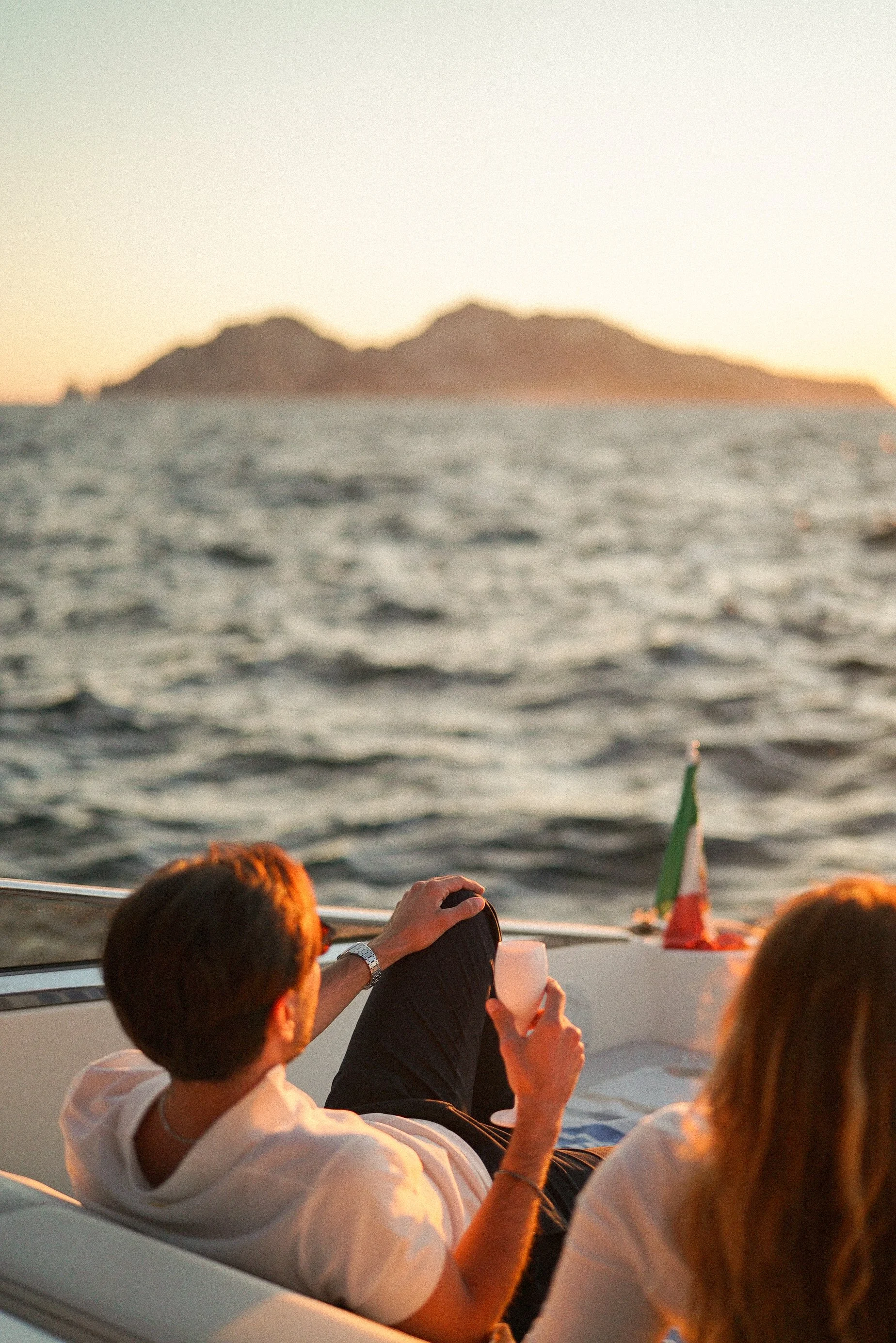 People relaxing on a boat at sunset with mountains in the background, featuring a small Italian flag and a person taking a photo.