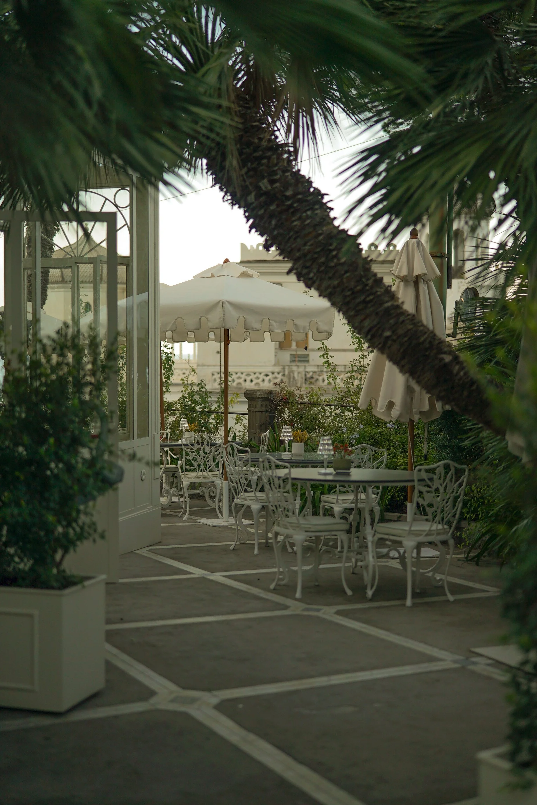 Outdoor patio area with white wrought iron tables and chairs, large white umbrellas, and lush greenery, including palm trees and potted plants.