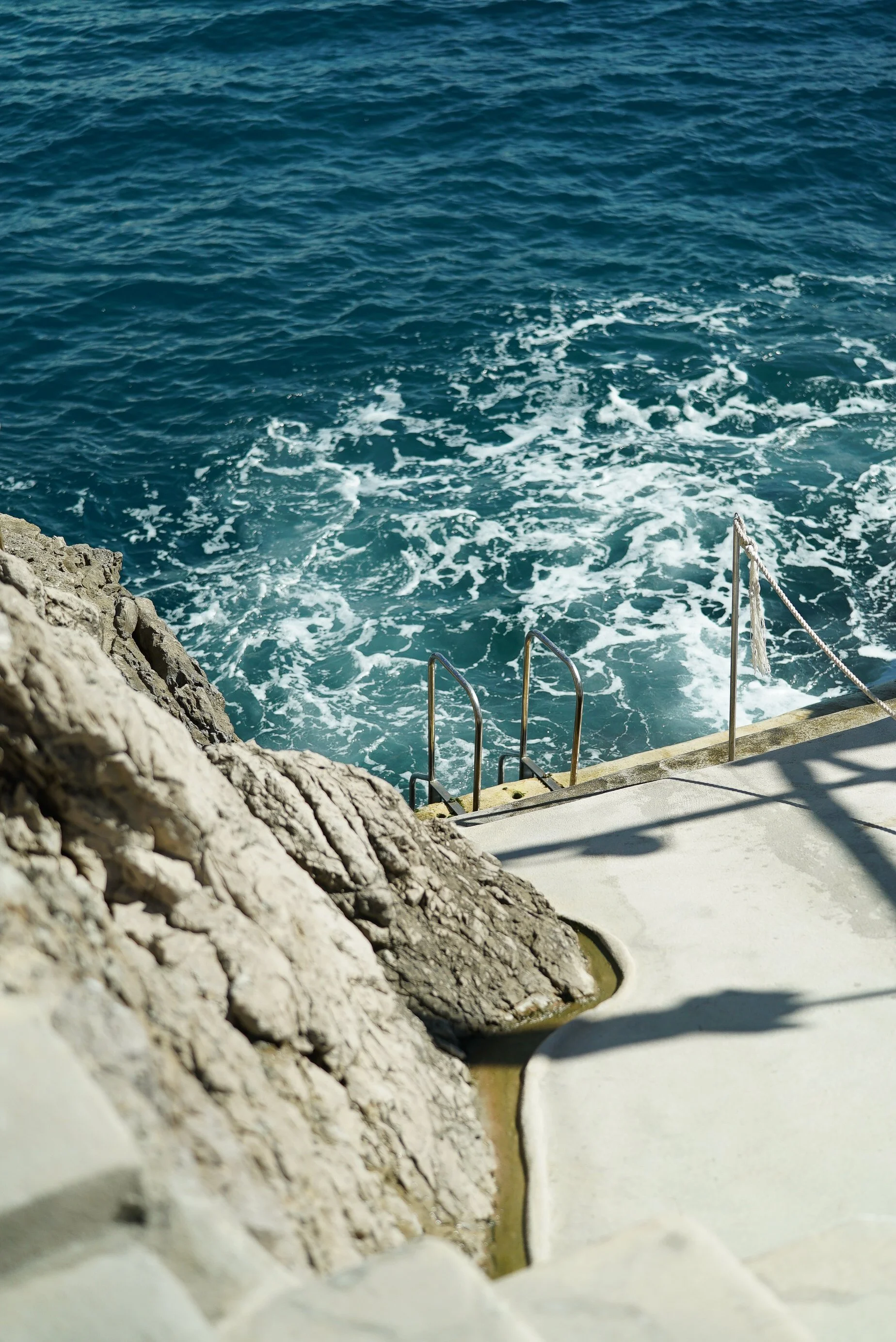 View of the ocean with blue water and waves near a rocky shore, seen from a boat or dock with white concrete steps and metal handrails.