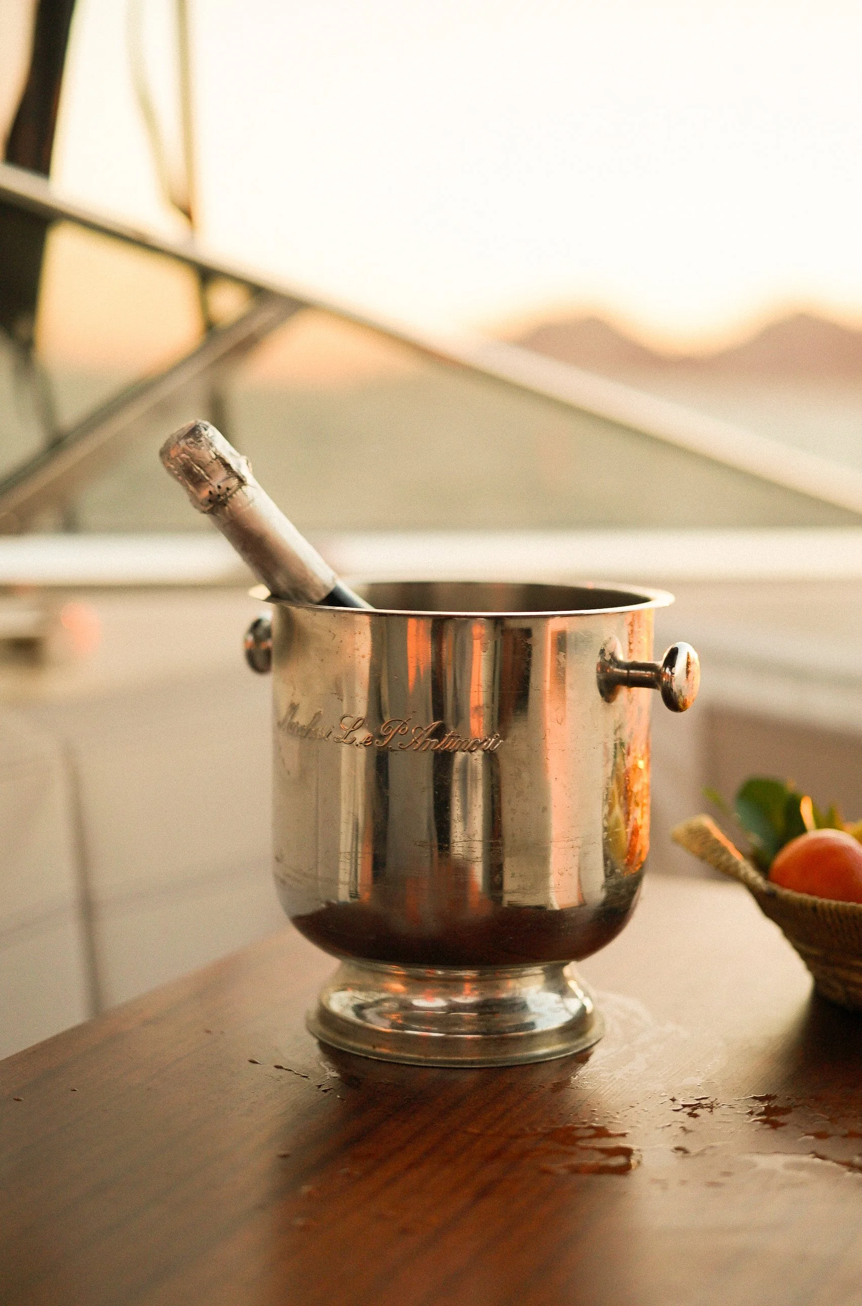 A silver trophy cup with a handle and engraved text, placed on a wooden surface with spilled wine around it. The background is blurred with a warm, soft light.