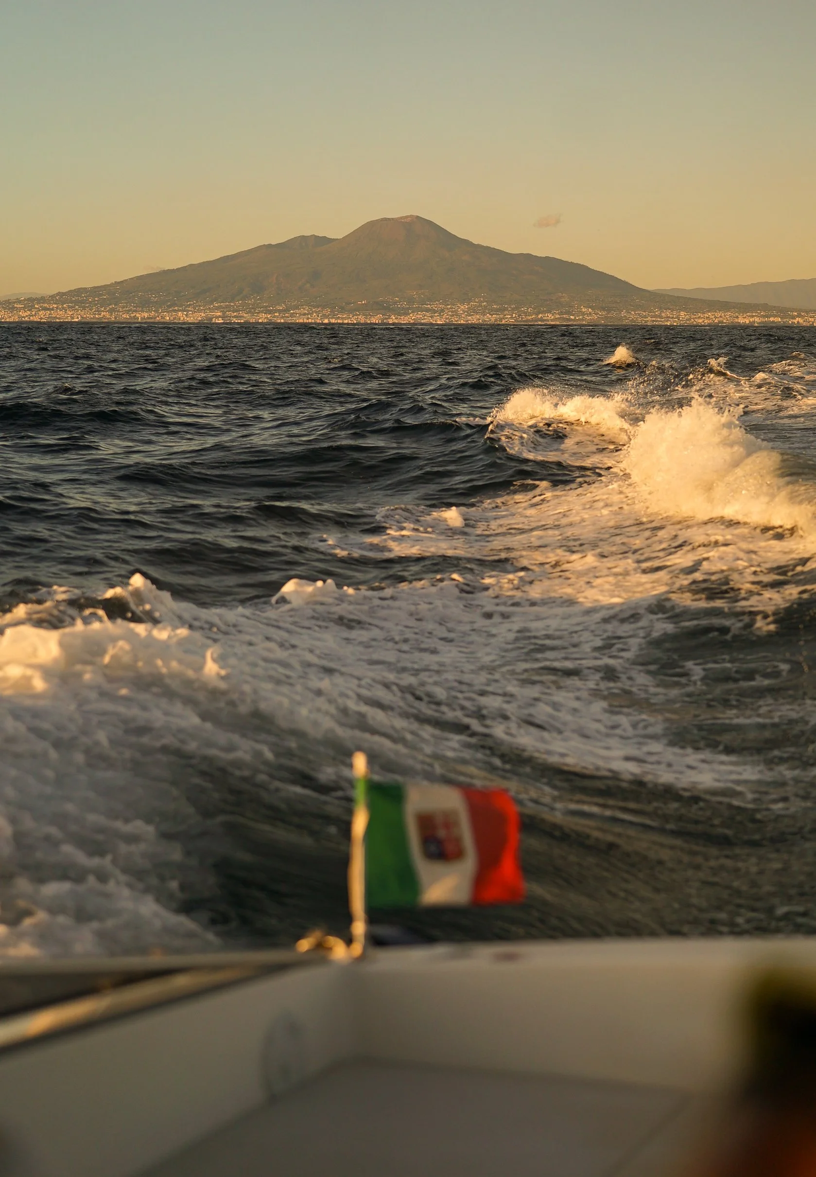 View from a boat showing the sea, with a small Italian flag in the foreground, and a large mountain or volcano in the distance during sunset or sunrise.
