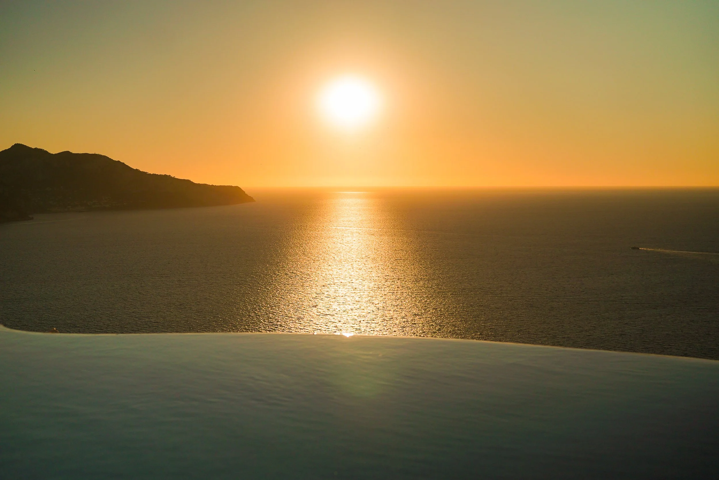 Sun setting over the ocean with a silhouette of a landmass on the left side and a boat cruising on the water.