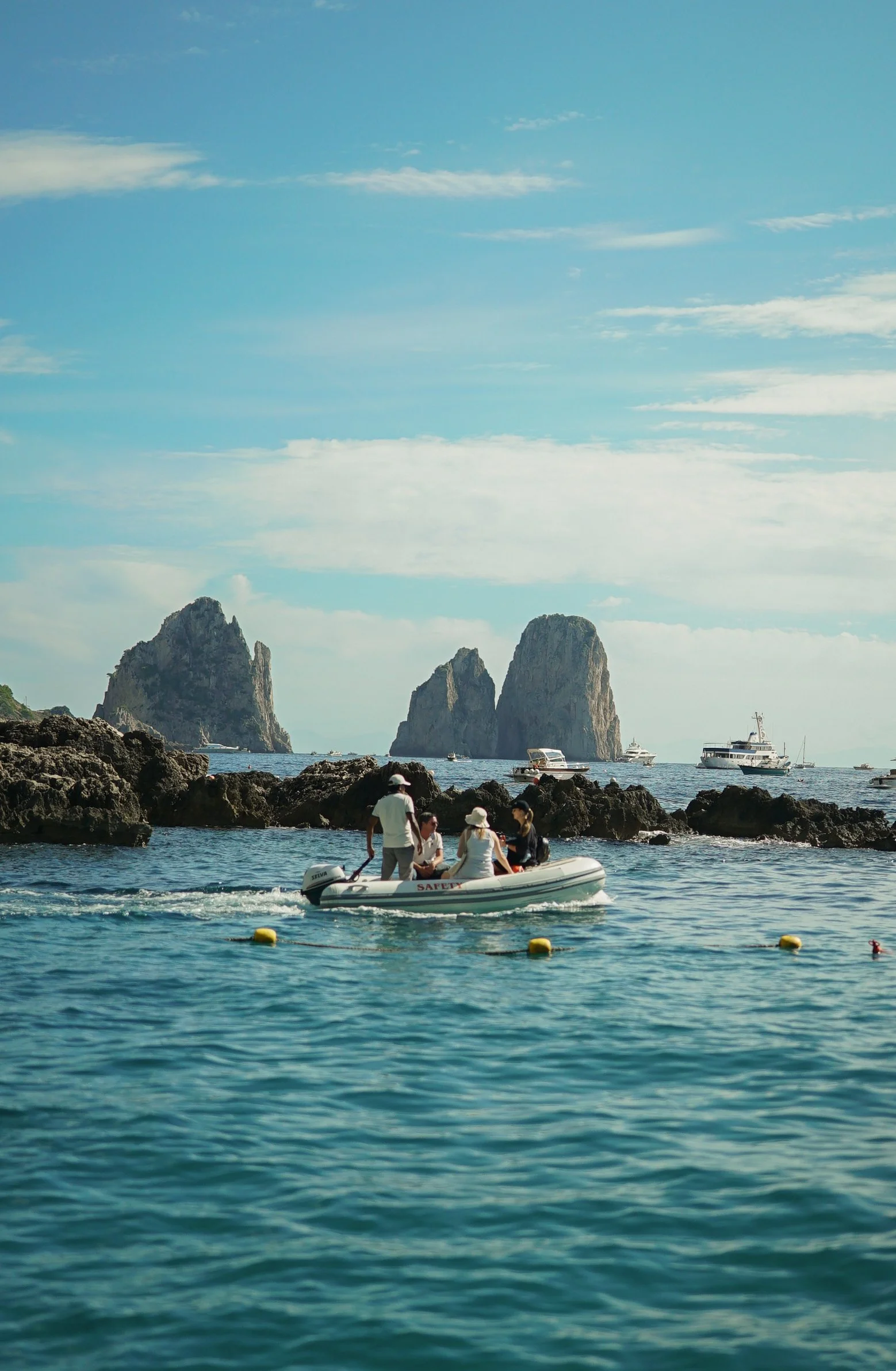 People riding a small inflatable boat in the ocean near rocky islands and a coastline under a partly cloudy sky.