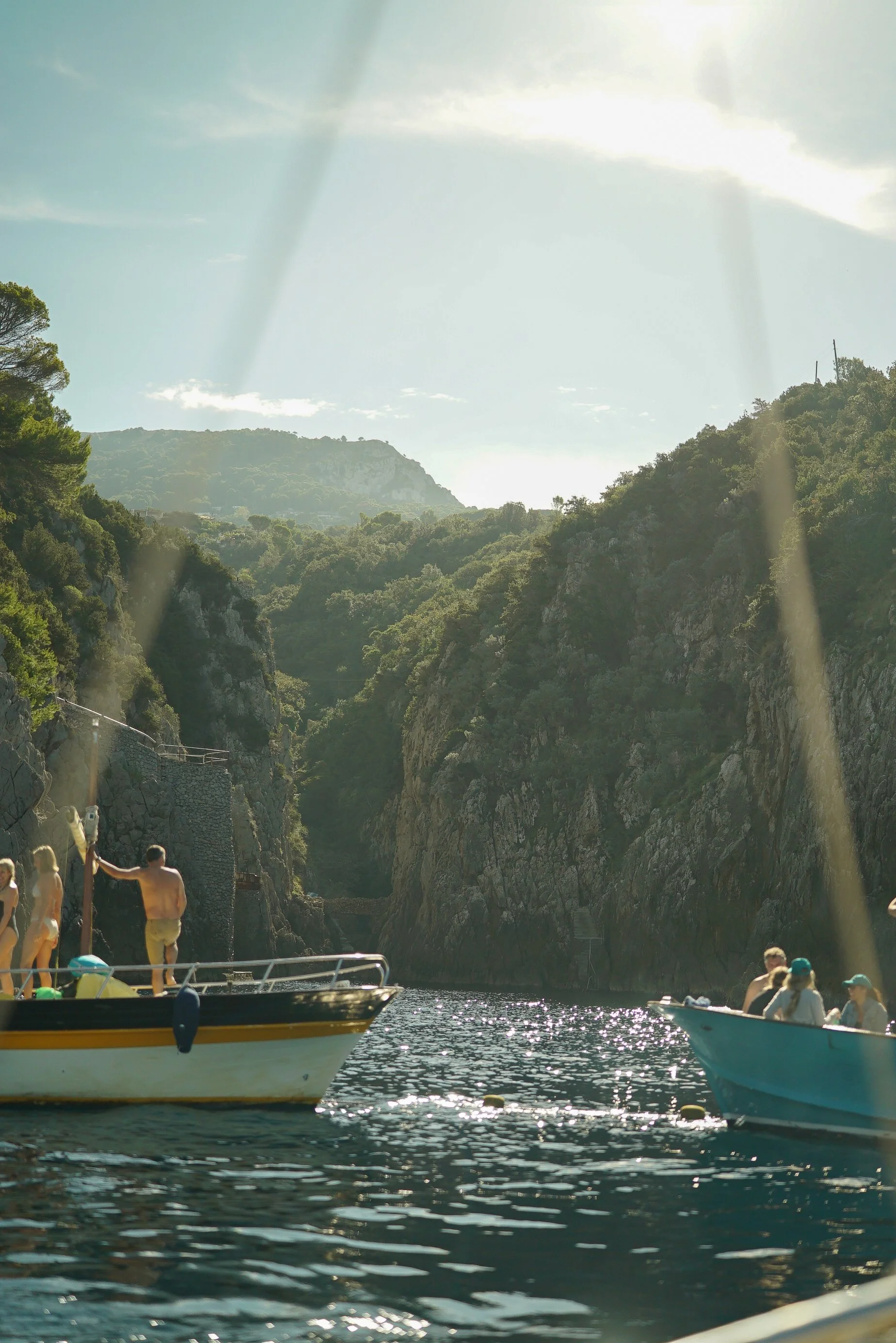 People in swimwear on boats in a scenic canyon with cliffs and lush greenery, sunlight streaming through clouds.