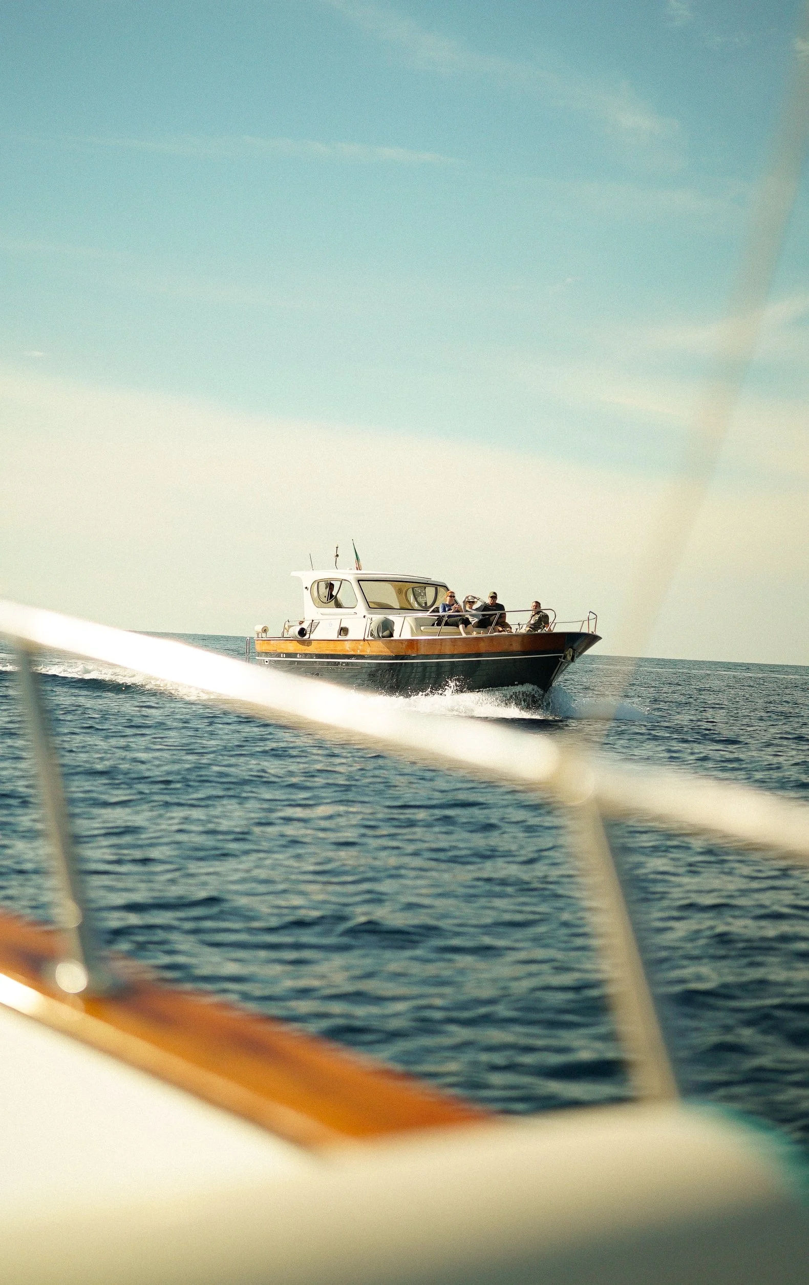 A small boat with a white cabin and wooden trim sailing on the water with four people aboard. The photo is taken from another boat, with part of its railing and a blurred part of the boat in the foreground. Clear, calm ocean under a blue sky.