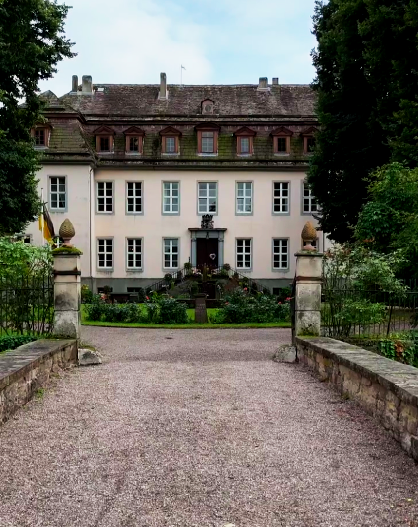 Ein großes, historisches Gebäude mit mehreren Fensterreihen und einem zentralen Eingang, umgeben von einem Garten mit Wegen und Zäunen, im Vordergrund ein gepflasterter Weg. Es handelt sich um das Rittergut in Meinbrexen.