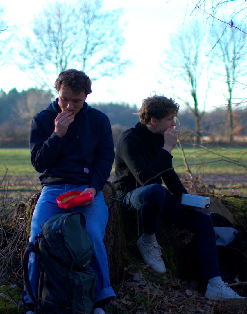 Zwei junge Männer, Fynn Jacob Utermark und Anton von Mansberg, sitzen auf einem Baumstamm im Freien und essen. Es ist Winter oder Frühling, Bäume sind kahl, im Hintergrund ein offenes Feld und Bäume.